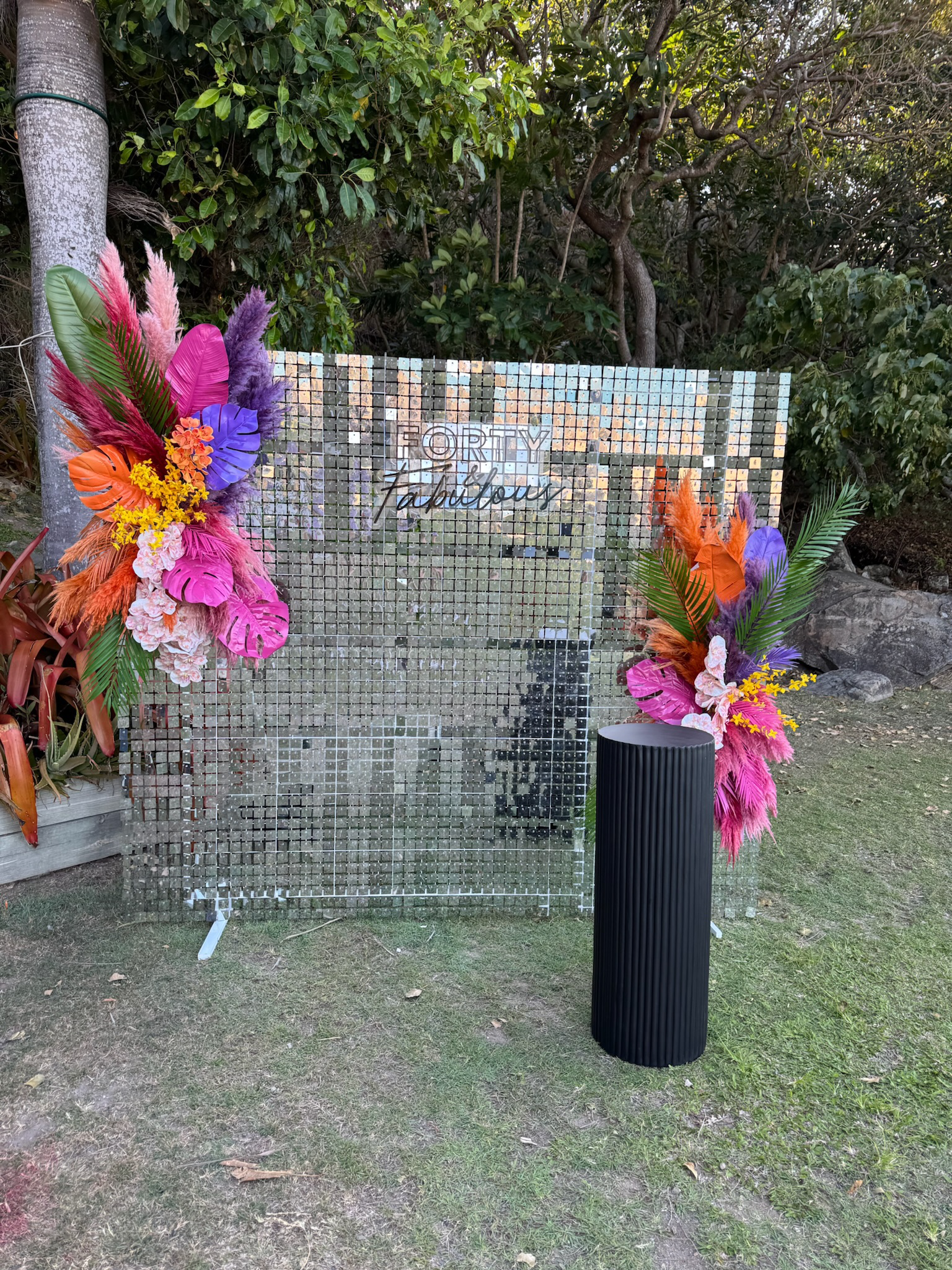 A decorative outdoor display features a mirrored mosaic backdrop with the words 'Very fabulous' written on it, surrounded by vibrant floral arrangements with pink, purple, orange, yellow, and green feathers and leaves, set on a grassy area with trees