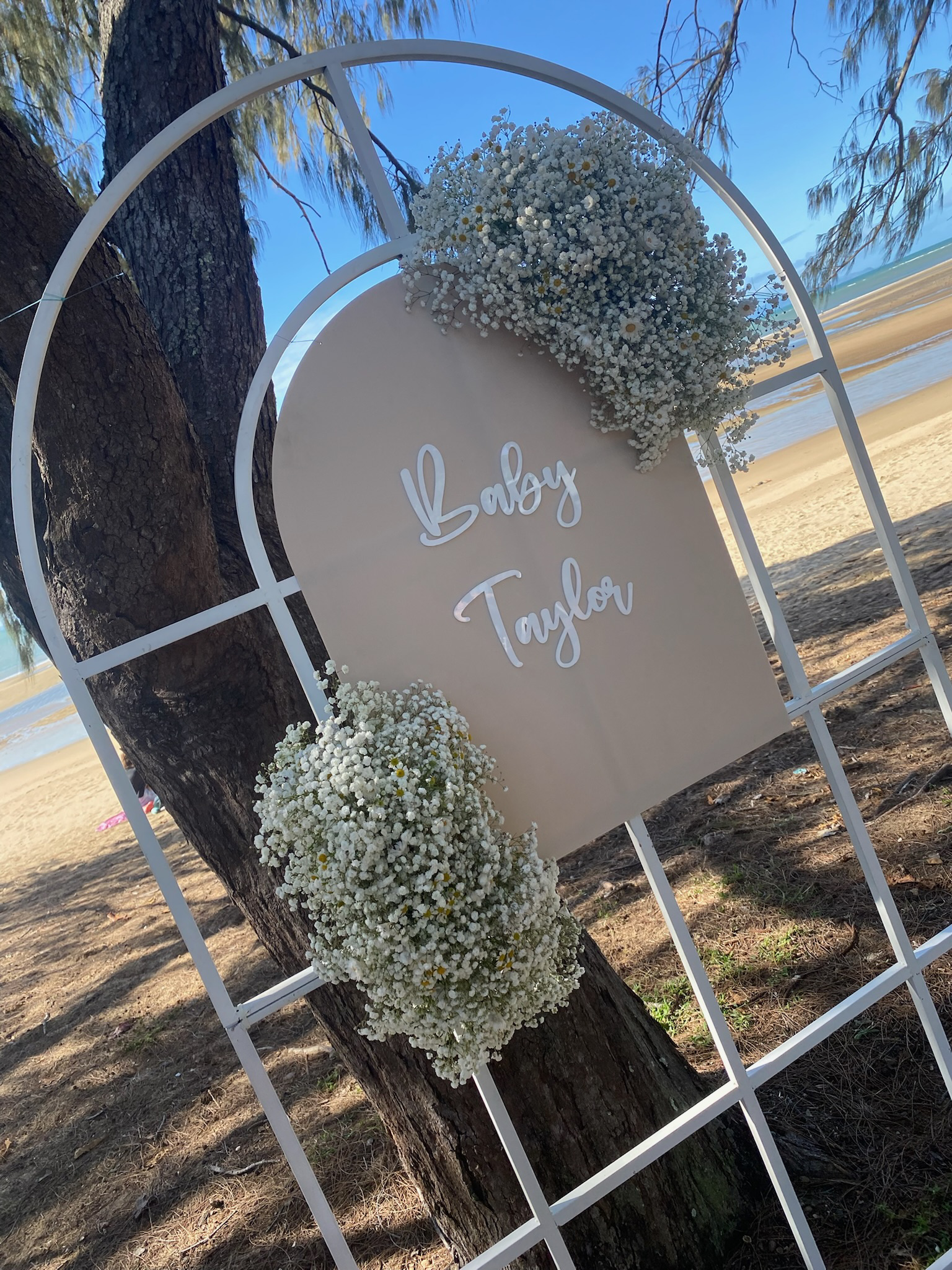A decorative sign with the words 'Baby Taylor' written on it, surrounded by white flower arrangements, placed outdoors near a beach and trees.