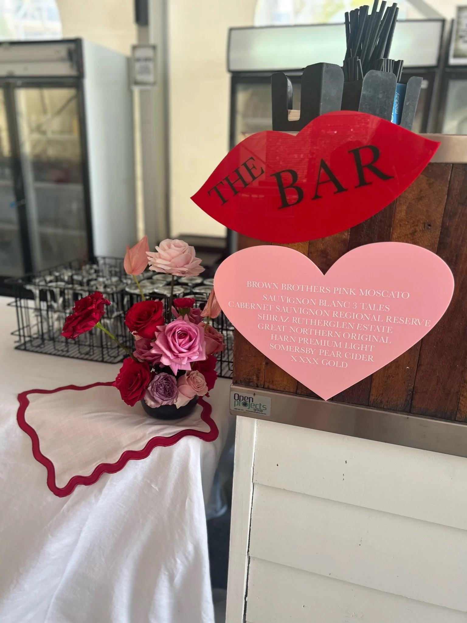 A Valentine-themed bar sign with a red lips graphic and a pink heart-shaped menu listing wine options, next to a small bouquet of pink and red roses on a table covered with a white cloth and red trim.