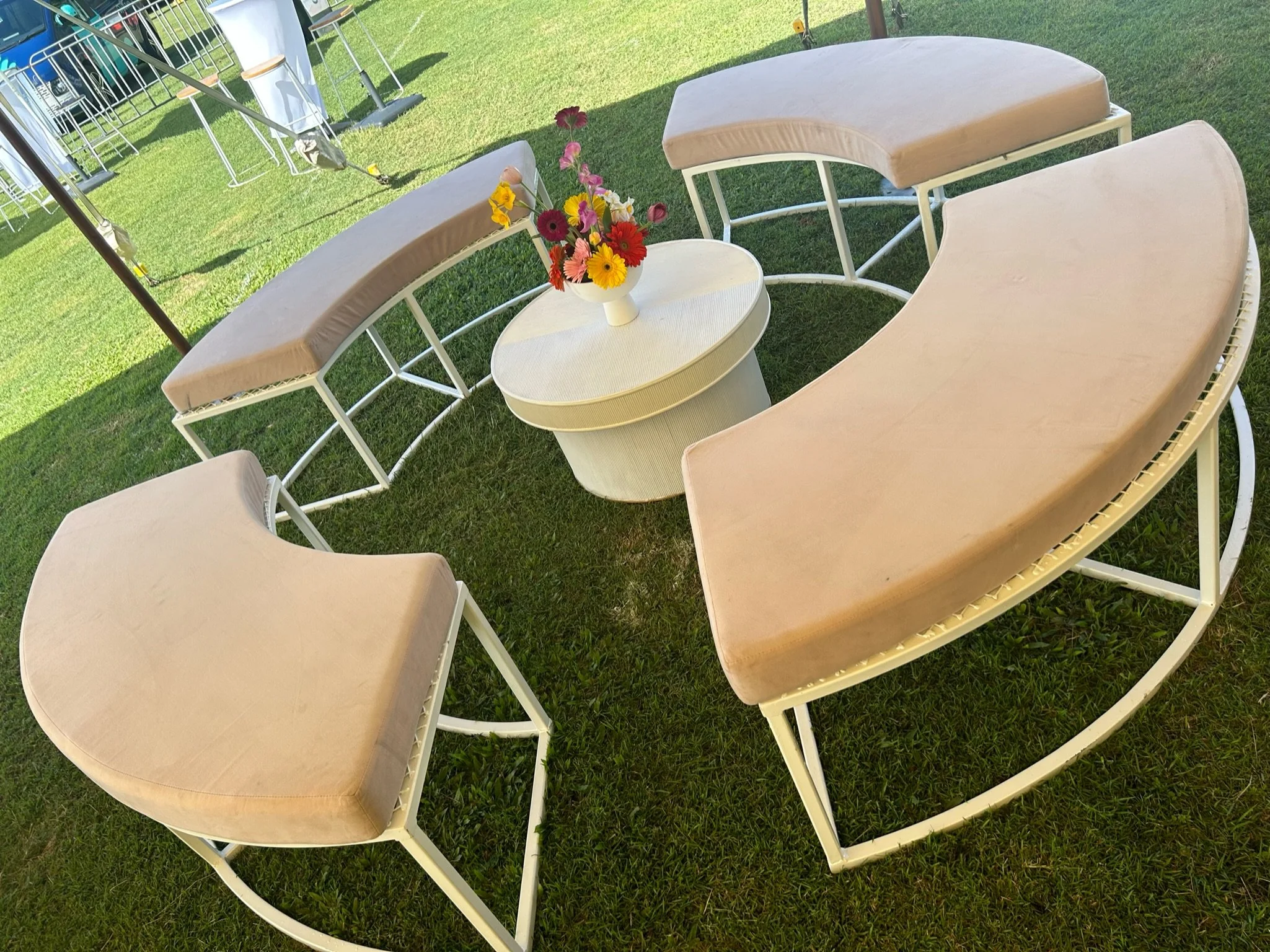 Outdoor seating area with curved beige cushioned benches and a small round white table holding a colorful flower arrangement, set on green grass.
