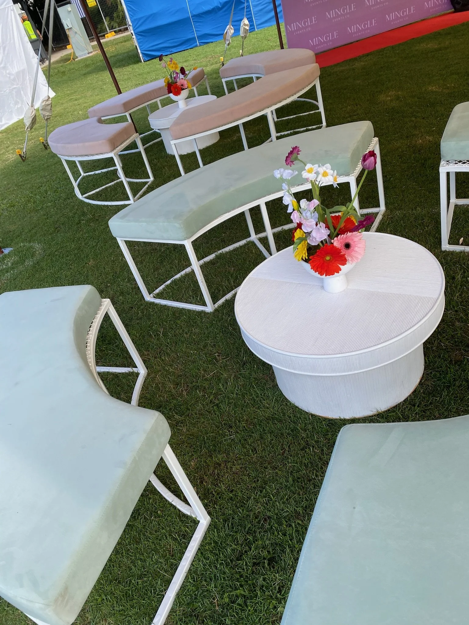Outdoor seating area with pastel-colored benches and a round white table with a flower arrangement in a white vase, set on green grass.