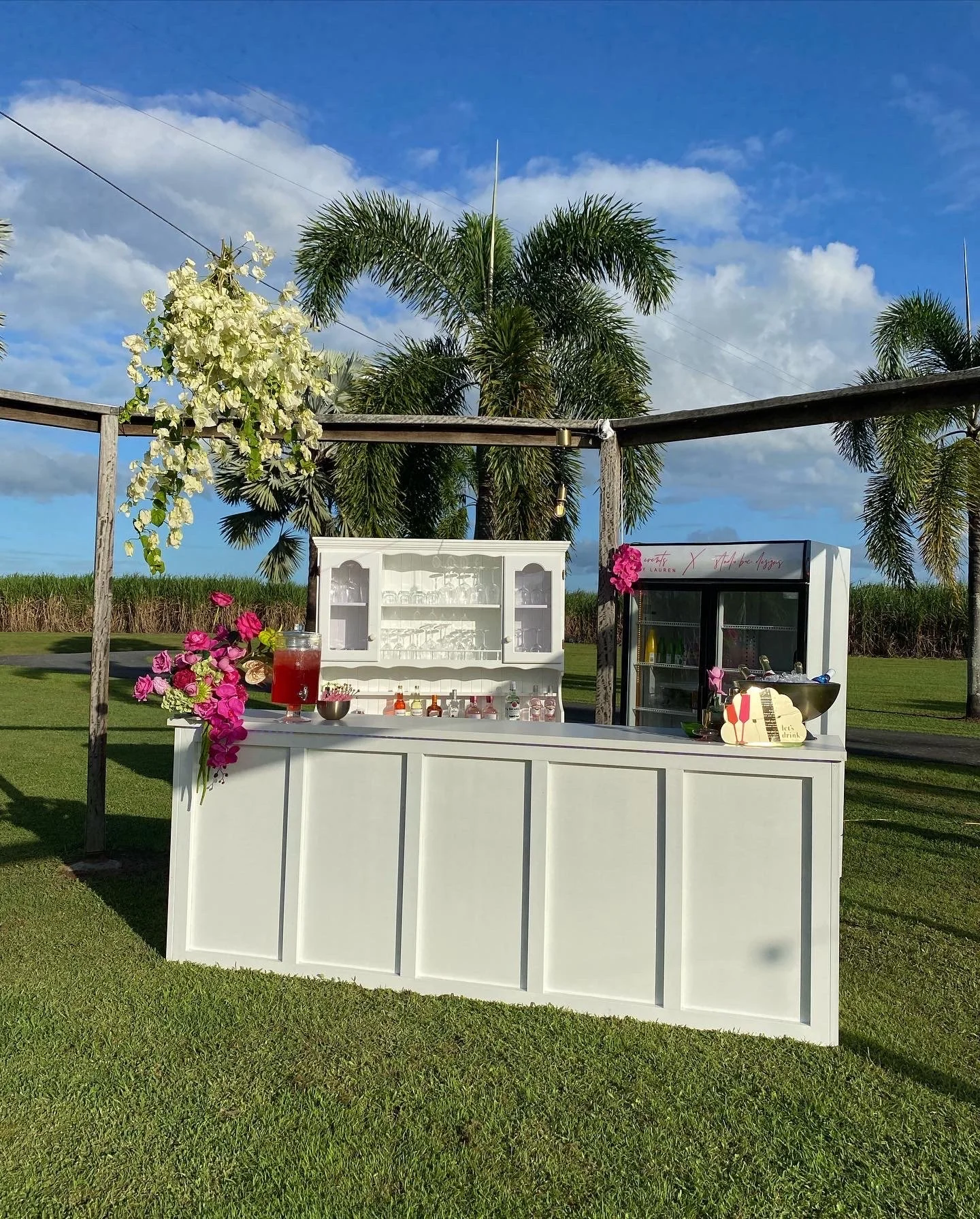 Outdoor refreshment stand with flowers, drinks, and glassware, set against palm trees and a blue sky.