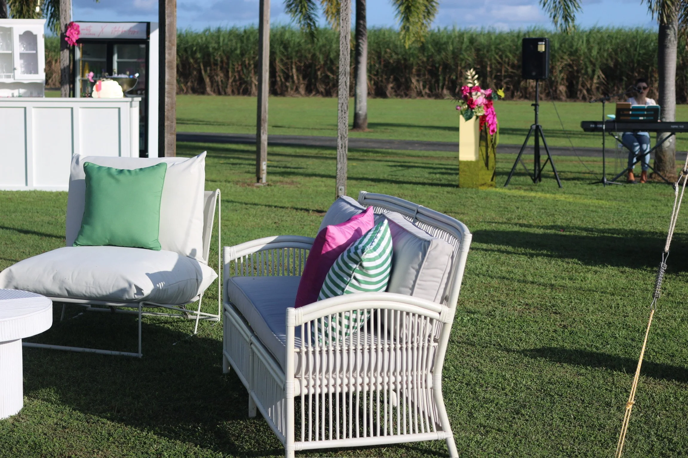 Outdoor event setup with white wicker and metal chairs with colorful pillows, a pink floral arrangement, and a musician playing keyboard in the background.