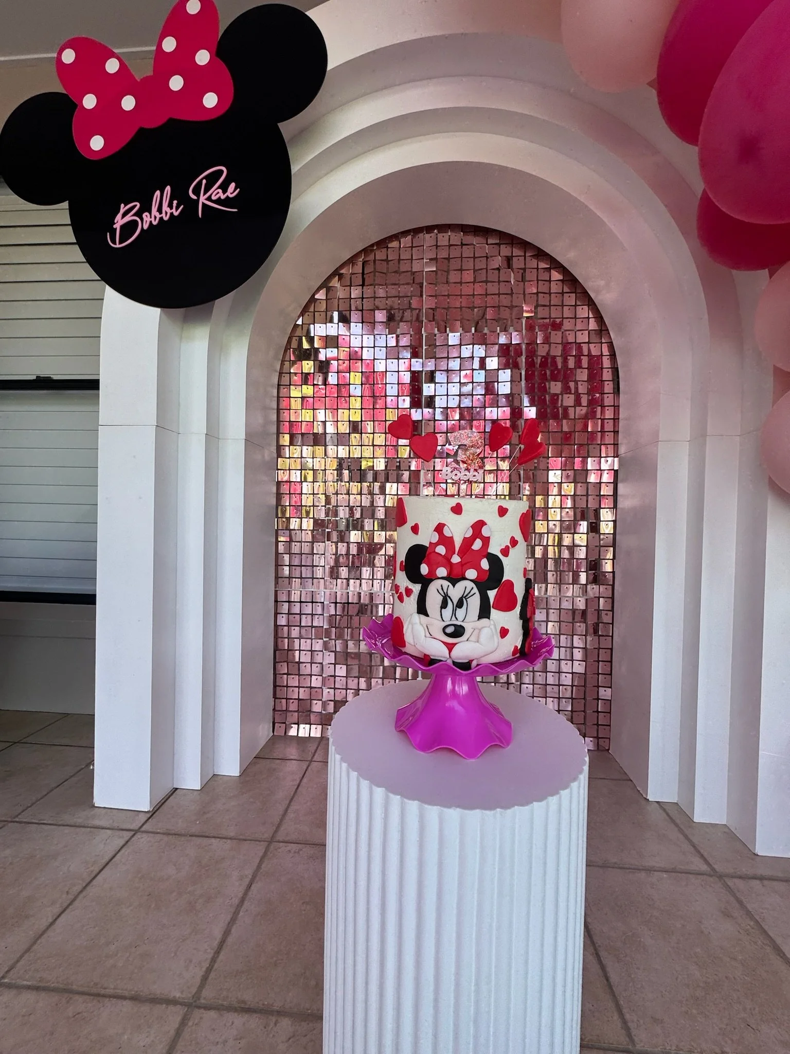 Minnie Mouse-themed birthday cake with Minnie’s face, red and white polka dot bow, and red hearts, displayed on a pink cake stand against a pink and rose gold backdrop with balloons and a Minnie Mouse sign.