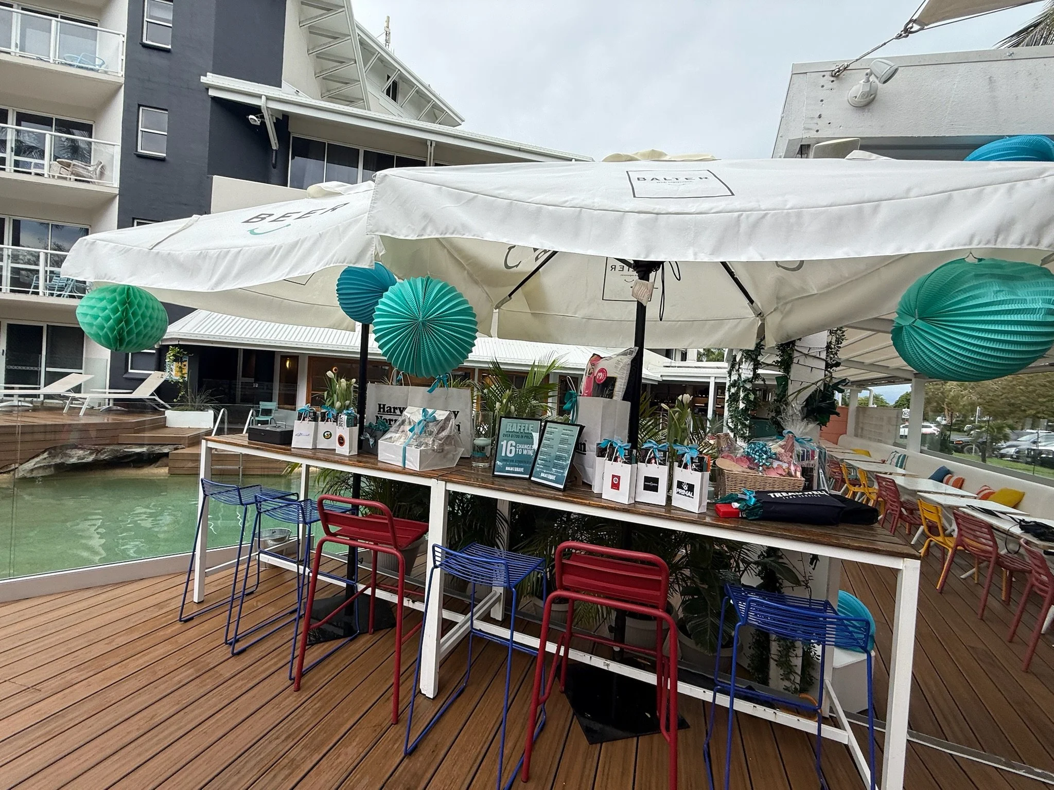 Outdoor poolside booth with a white canopy, blue and green paper lanterns, and a table with raffle prizes and gift bags, surrounded by colorful chairs on a wooden deck.