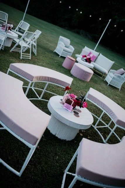 Outdoor garden party setup with white and pink furniture, including a round table with pink flowers, pink ottomans, and white chairs on a grassy lawn at dusk.