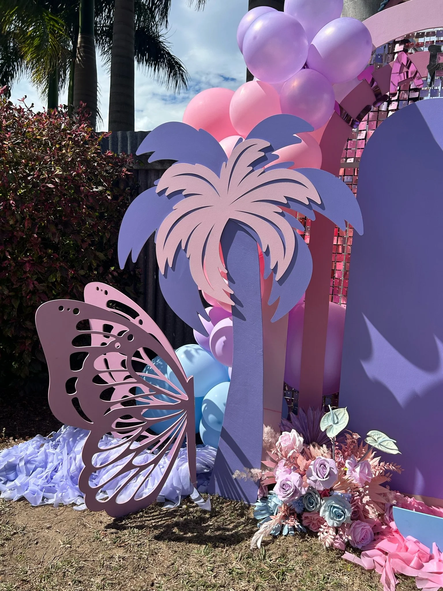 Decorative outdoor setup featuring pink and purple balloons, a pink and purple palm tree cutout, a lavender butterfly cutout, and a floral arrangement with roses and other flowers, with palm trees in the background and a partly cloudy sky.