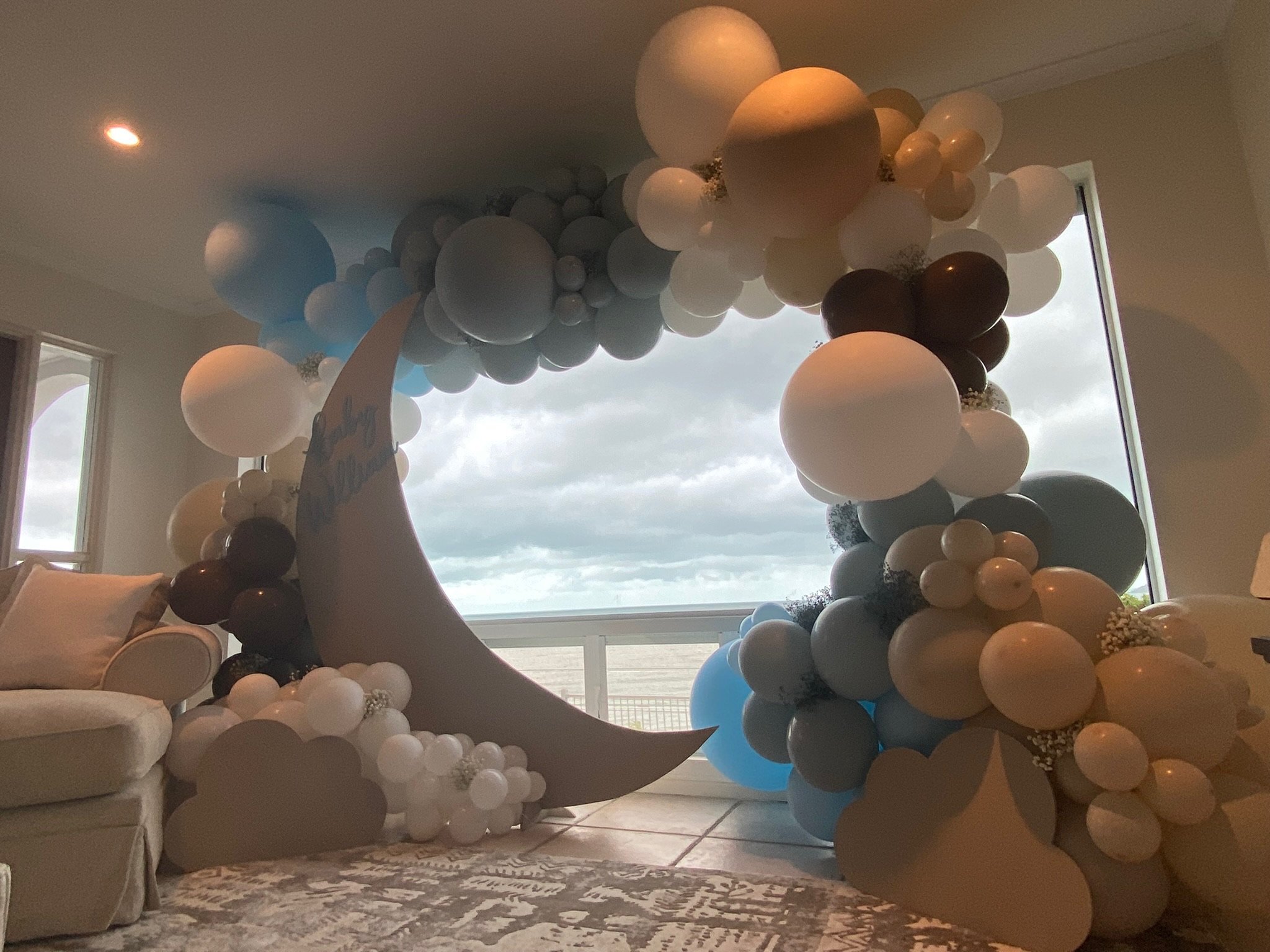 Interior of a room decorated with a large moon cutout and balloon arch in pastel colors, including white, brown, blue, and gray, placed near a window with cloudy sky outside.