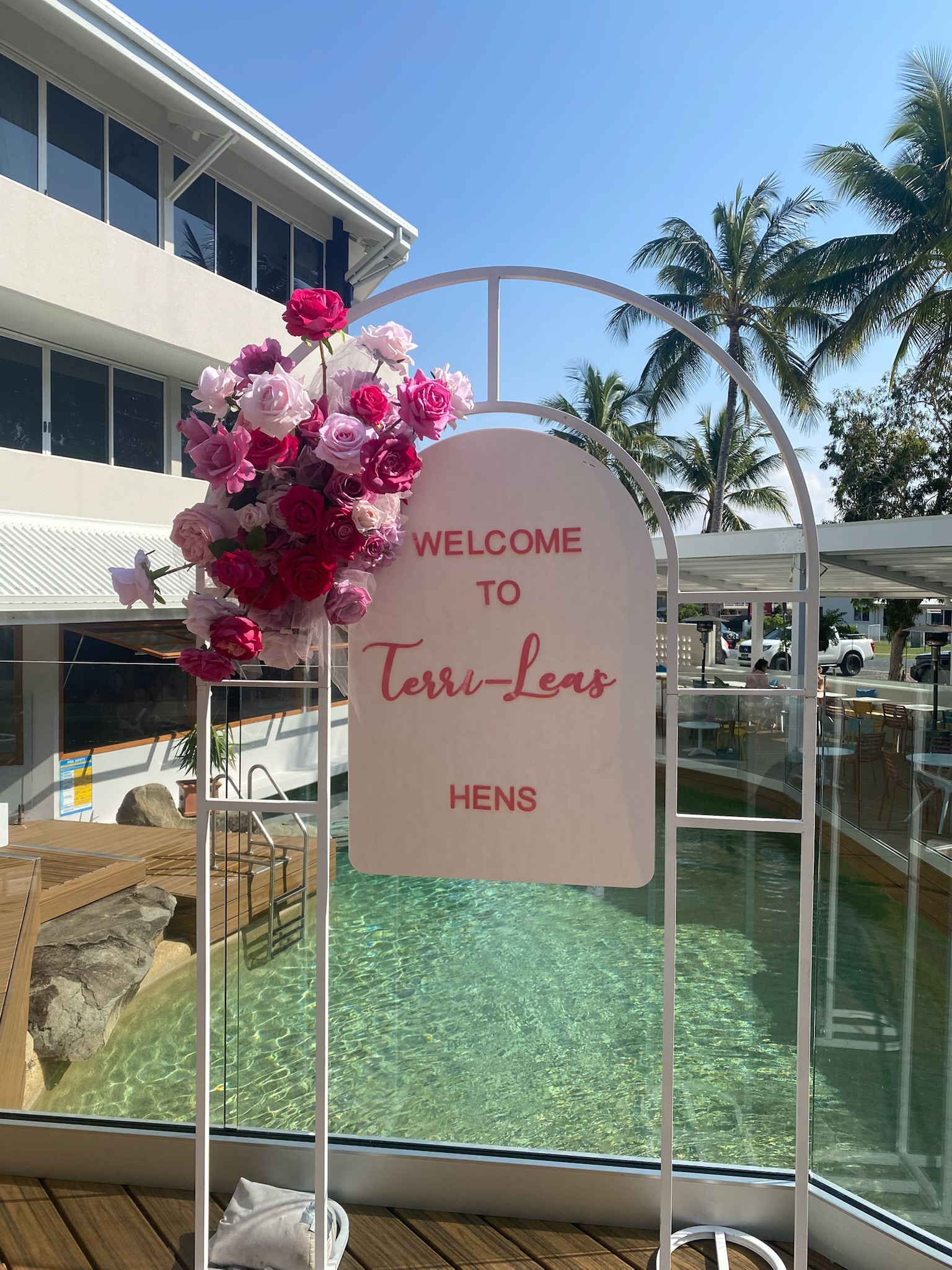 A welcoming sign for Terr-Leas Hens decorated with a bouquet of pink and red roses, situated near a pool with clear water and a sunny, palm-tree-lined background.