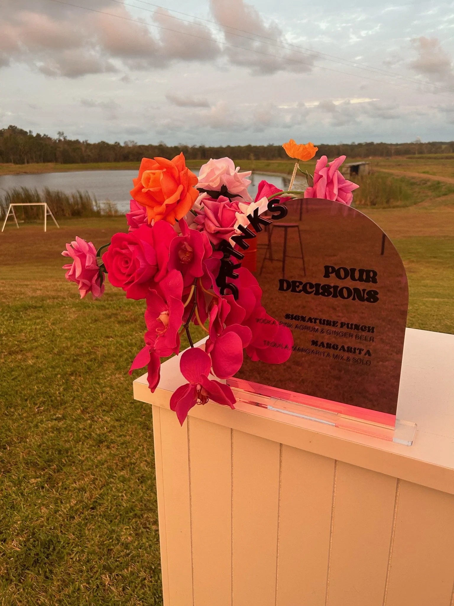 Sign with pink, red, orange, and purple flowers in front of a pond and open landscape under a cloudy sky.
