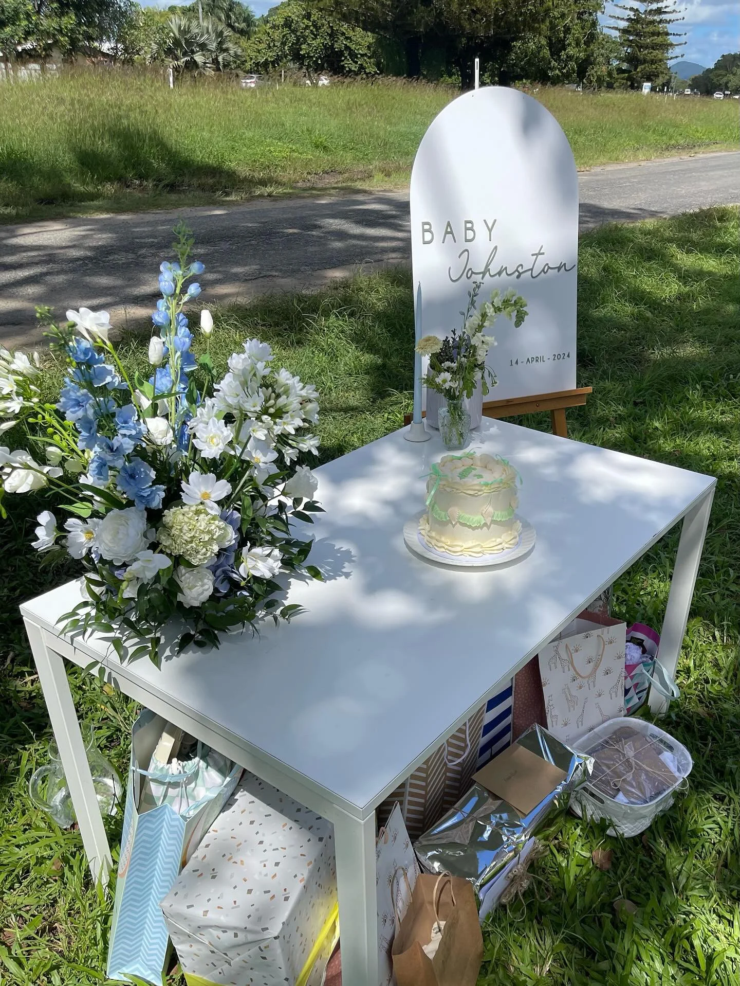 A small memorial setup outdoors with a white table, floral arrangements, a cake, and a sign that reads 'Baby Johnston 14 April 2024' in a grassy area near a roadside.