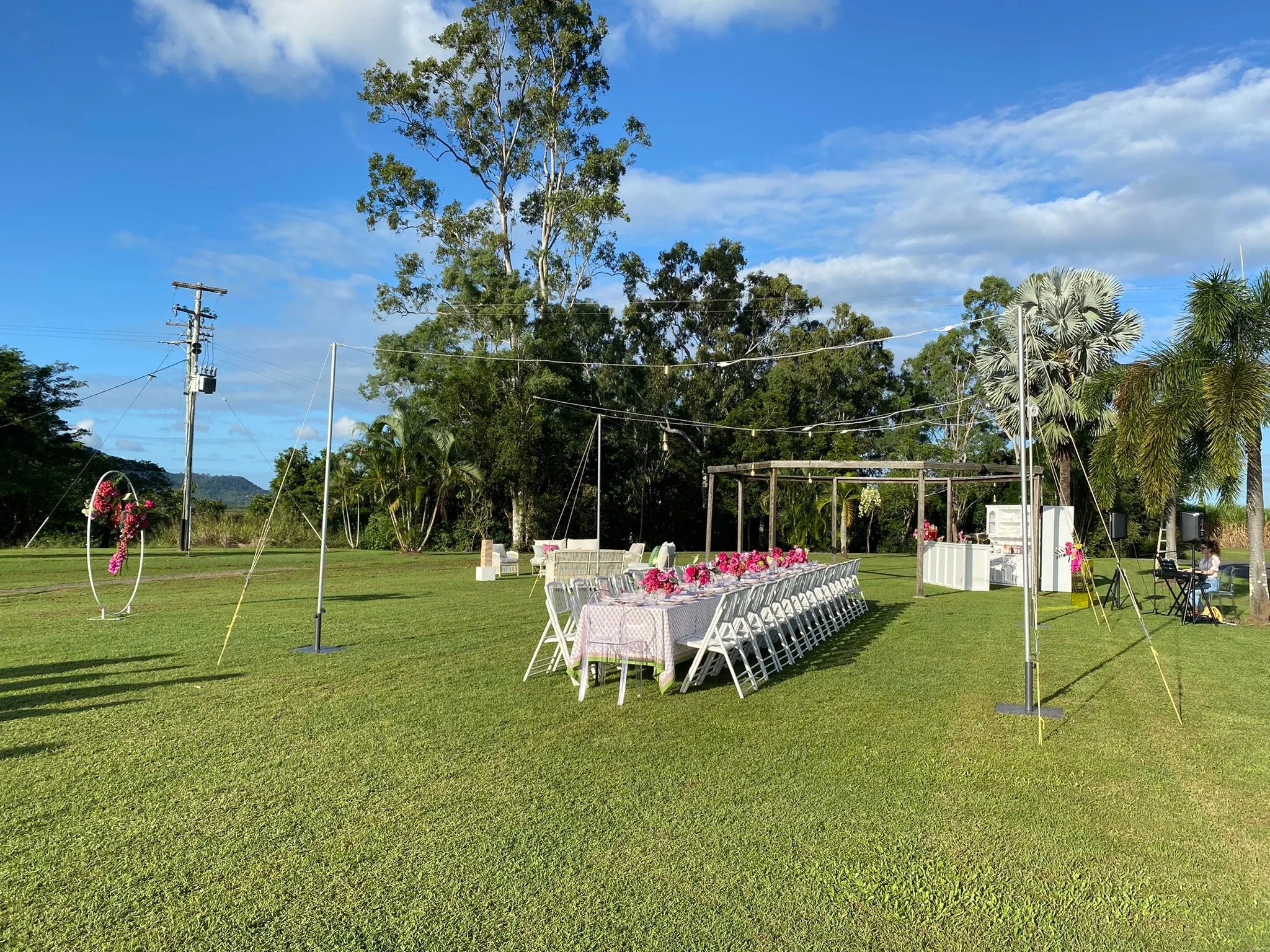 Outdoor event setup on a grassy field with a long table decorated with pink flowers, white chairs, a floral arch, and a small food and sound station with a musician playing keyboard nearby, under a blue sky with some clouds.