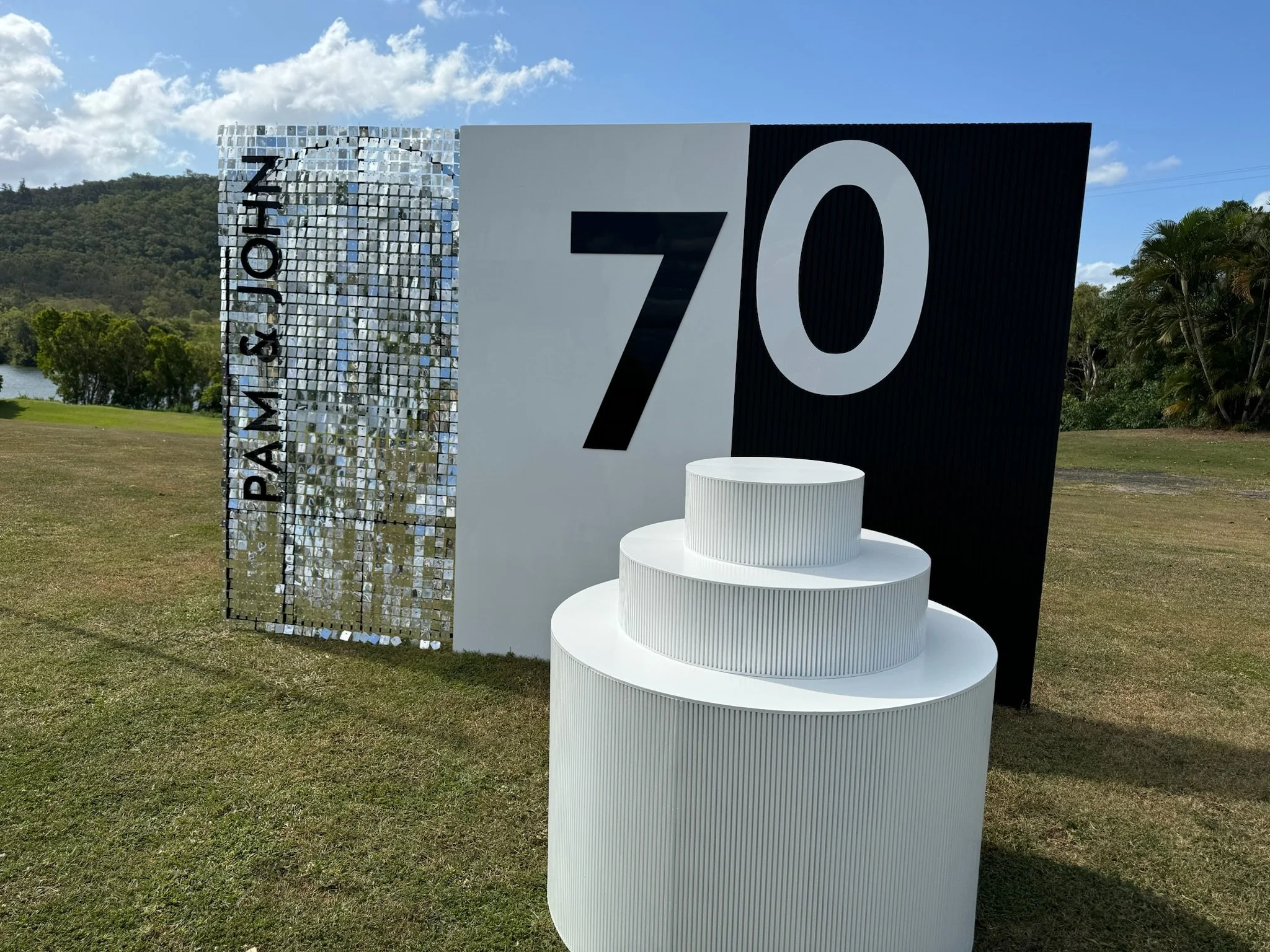 Sign with text indicating '70' and 'PAR-3 & JOHN' next to a three-tiered white podium on a grassy area outdoors, with trees and a body of water in the background under a partly cloudy blue sky.