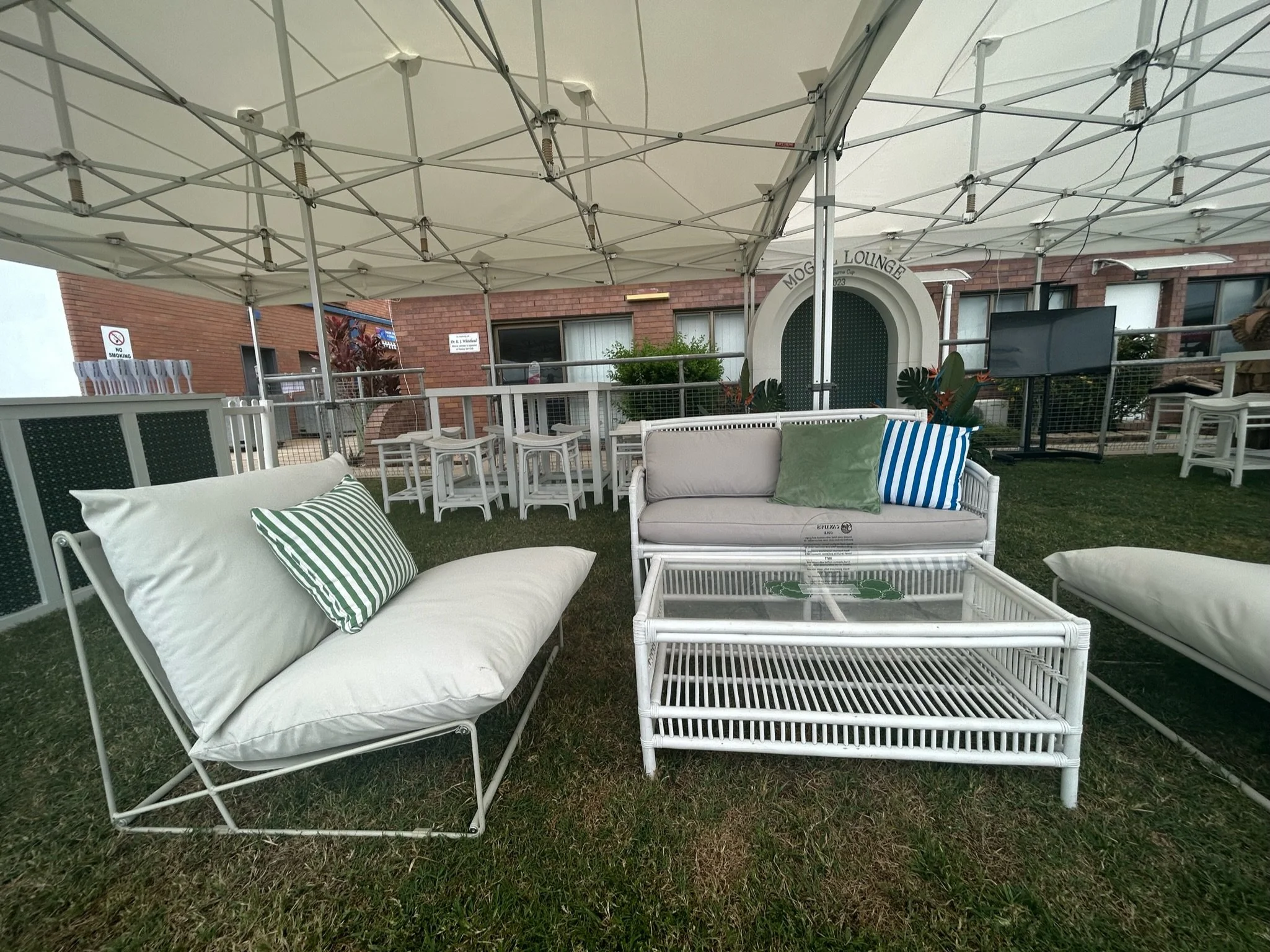 Outdoor seating area with white cushioned couches, striped and solid pillows, glass-top coffee table, under a large beige canopy, surrounded by artificial grass and brick building in the background.
