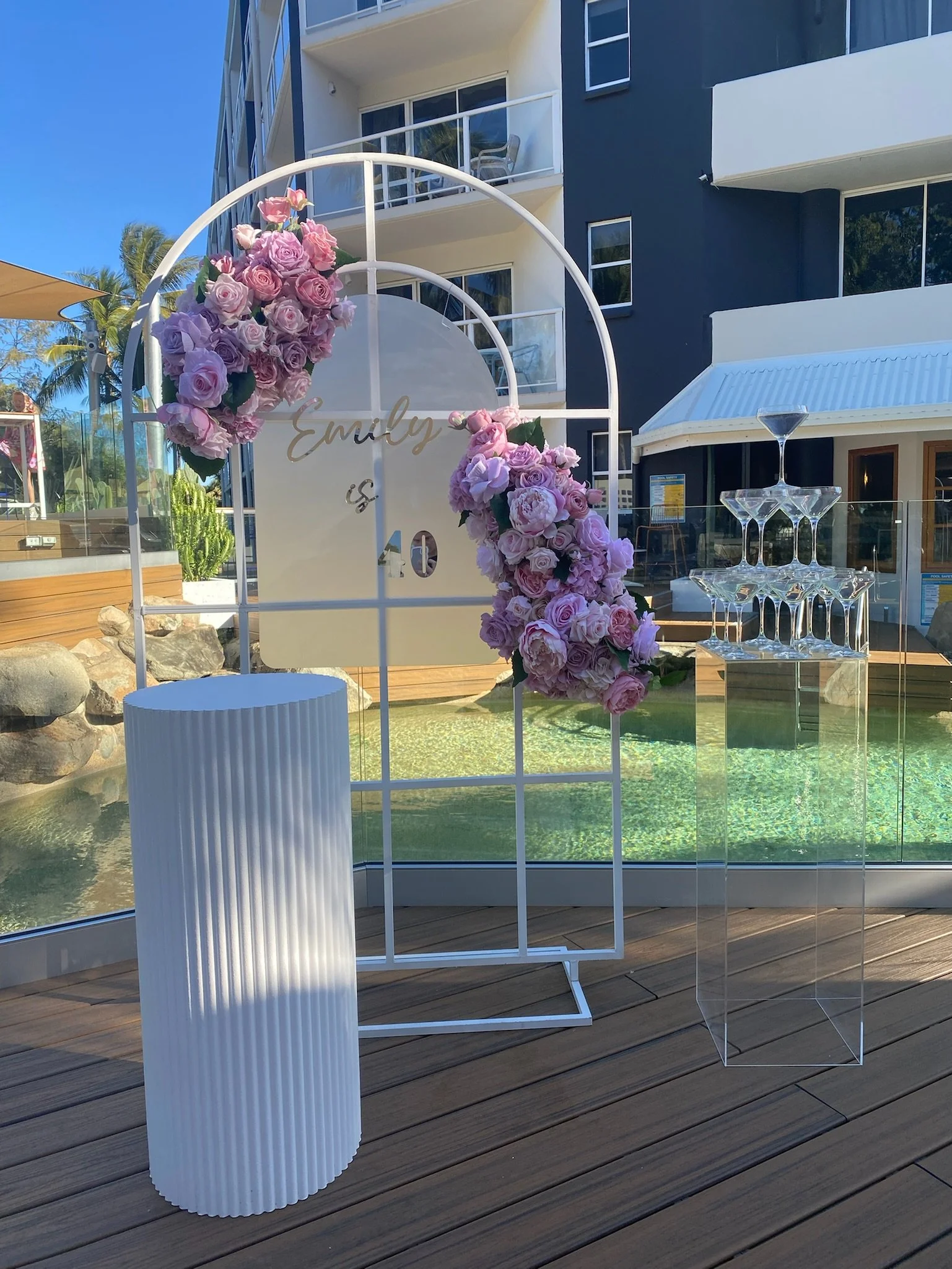 Decorative backdrop with pink and purple flowers, wedding sign reading 'Emely 10', stacking glassware on a clear acrylic stand, white pleated cylindrical table, and a swimming pool in the background at an outdoor event venue.