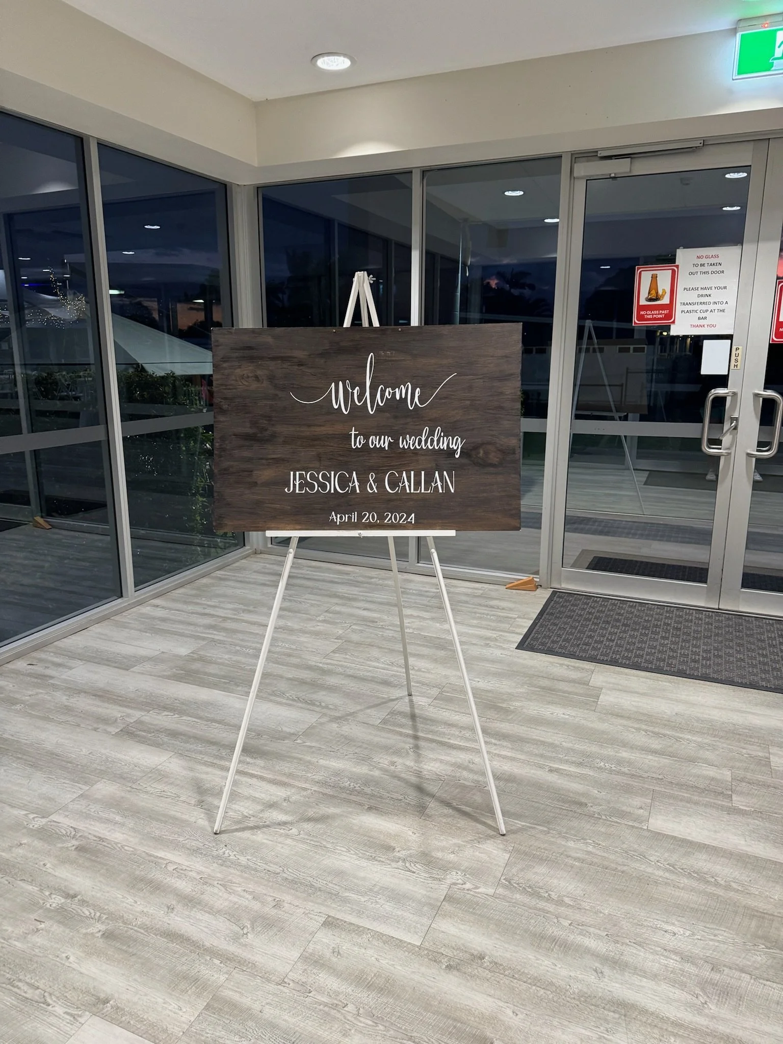 Wedding welcome sign on an easel that reads: Welcome to our wedding Jessica & Callan April 20, 2024, inside a building with glass doors and windows.