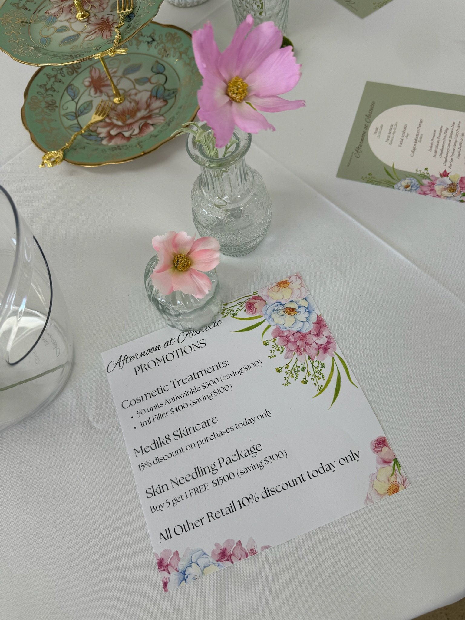 A table setting with floral dishware, pink flowers in glass vases, and a promotional flyer for beauty treatments.