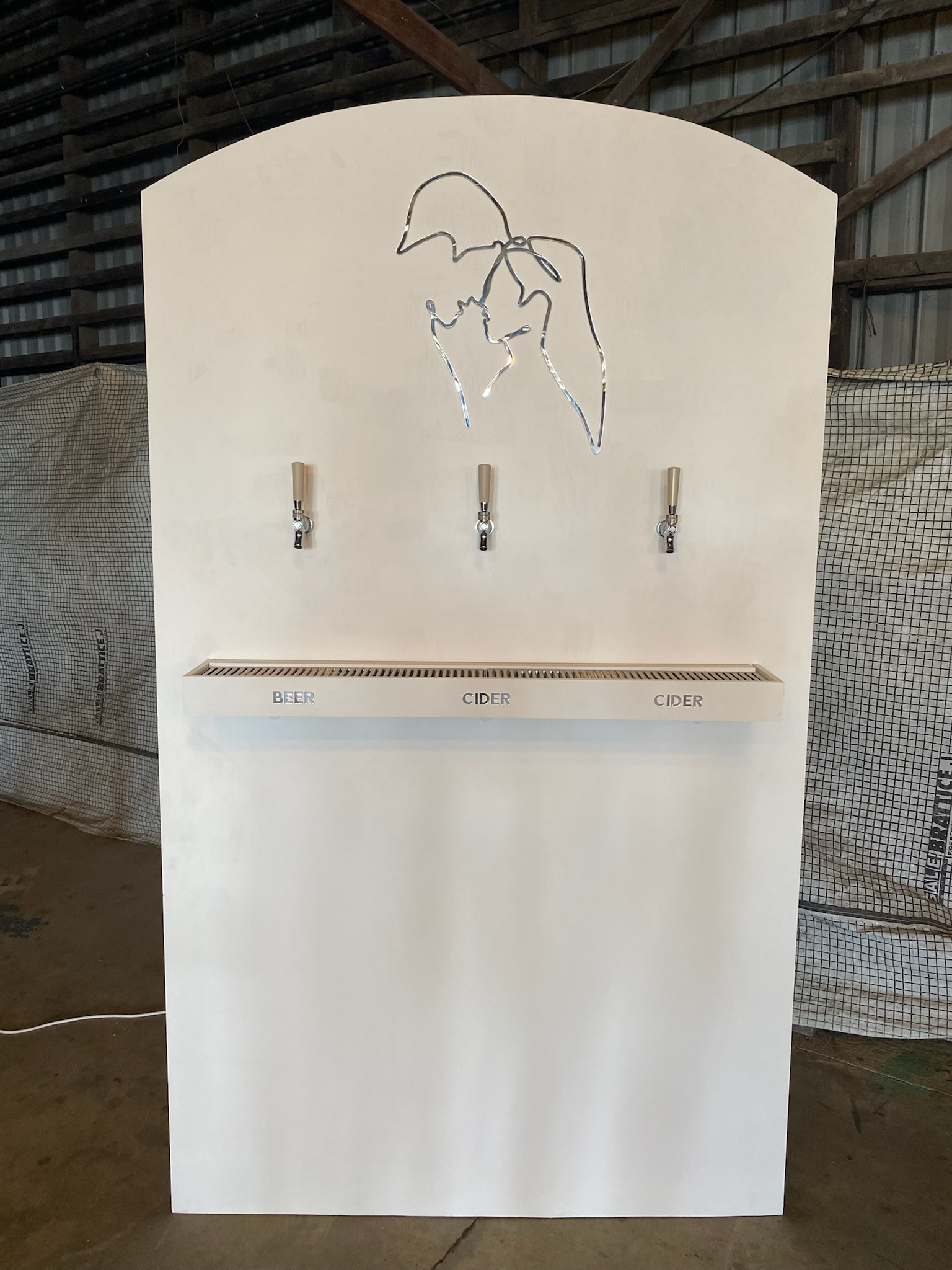 A white wall with a line art illustration of a person's face wearing a hat, mounted above three beer and cider taps. The taps are labeled "BEER," "CIDER," and "CIDER." There is a metal drain tray below the taps.