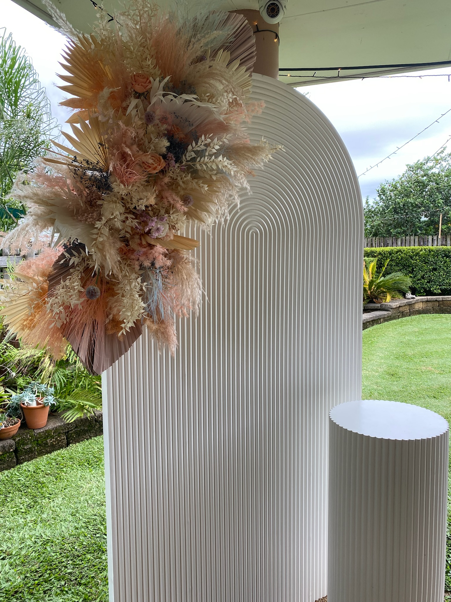 A decorative backdrop with an arch and a round pedestal, decorated with a large, cascading floral arrangement of dried and fresh flowers, pampas grass, and palms, set outdoors with a garden and cloudy sky in the background.