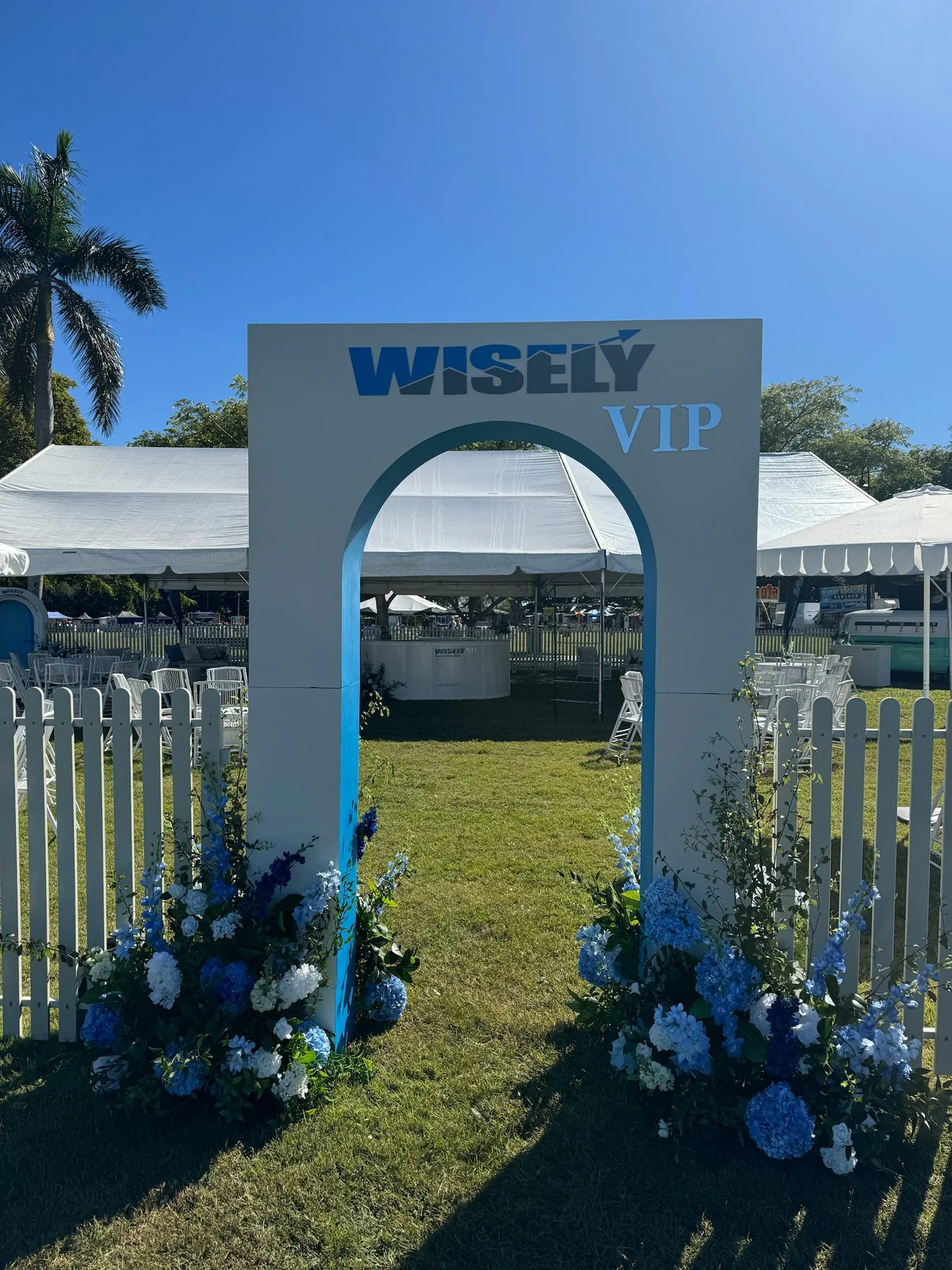 Entrance archway with 'WISELY VIP' sign, decorated with blue and white flowers, leading to an outdoor event area with white tents and seating, under a sunny, clear blue sky.