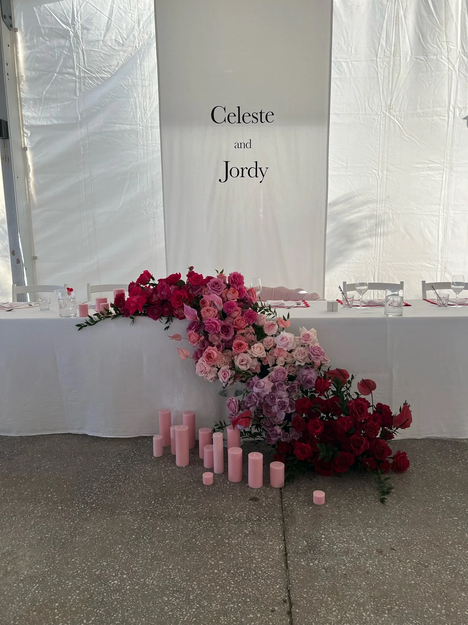 Wedding reception table decorated with a large pink and red flower arrangement and pink candles, with a backdrop displaying the names Celeste and Jordy.