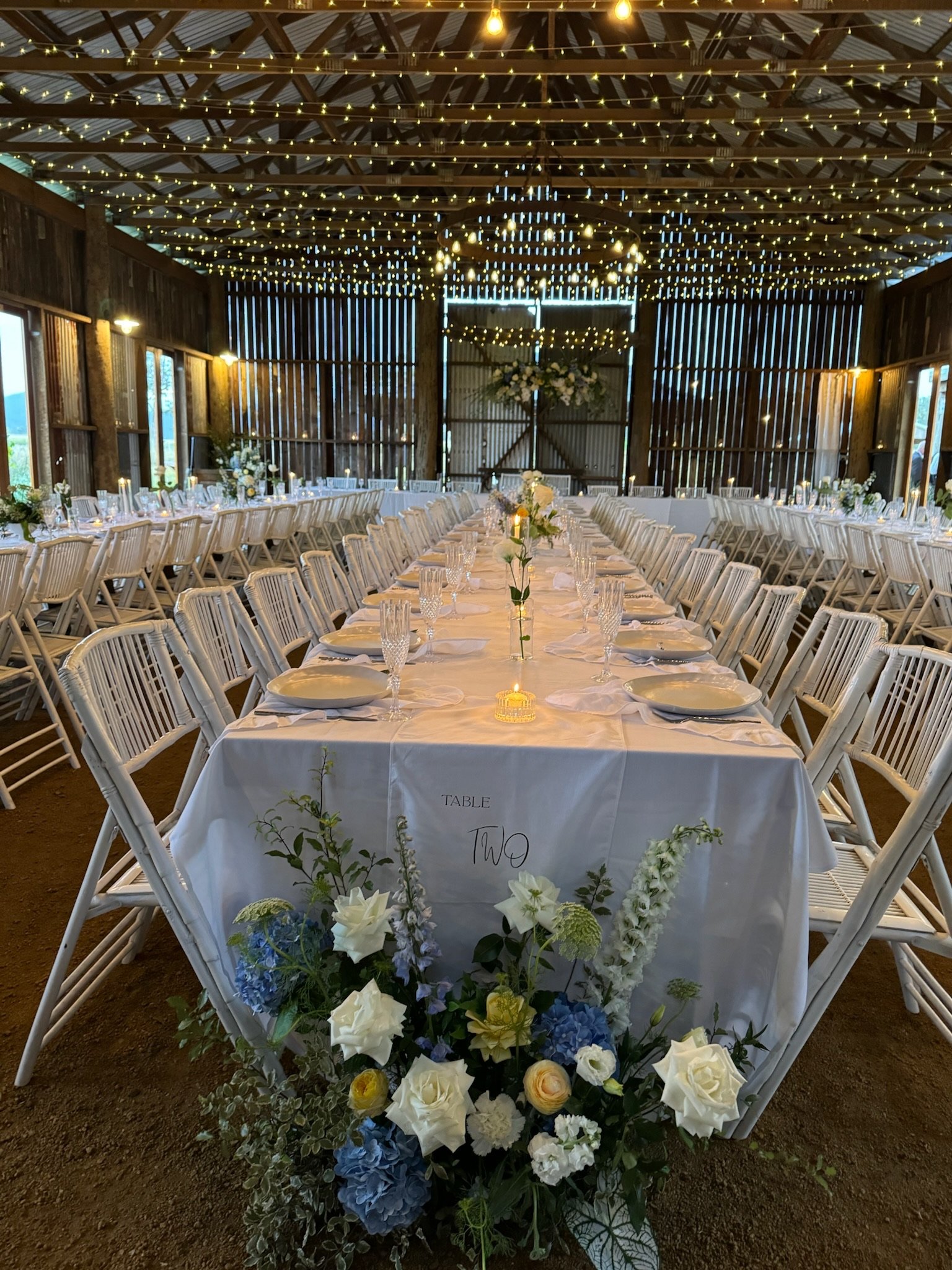 Wedding reception setup in barn with long tables, white chairs, decorative flowers, and string lights on ceiling.
