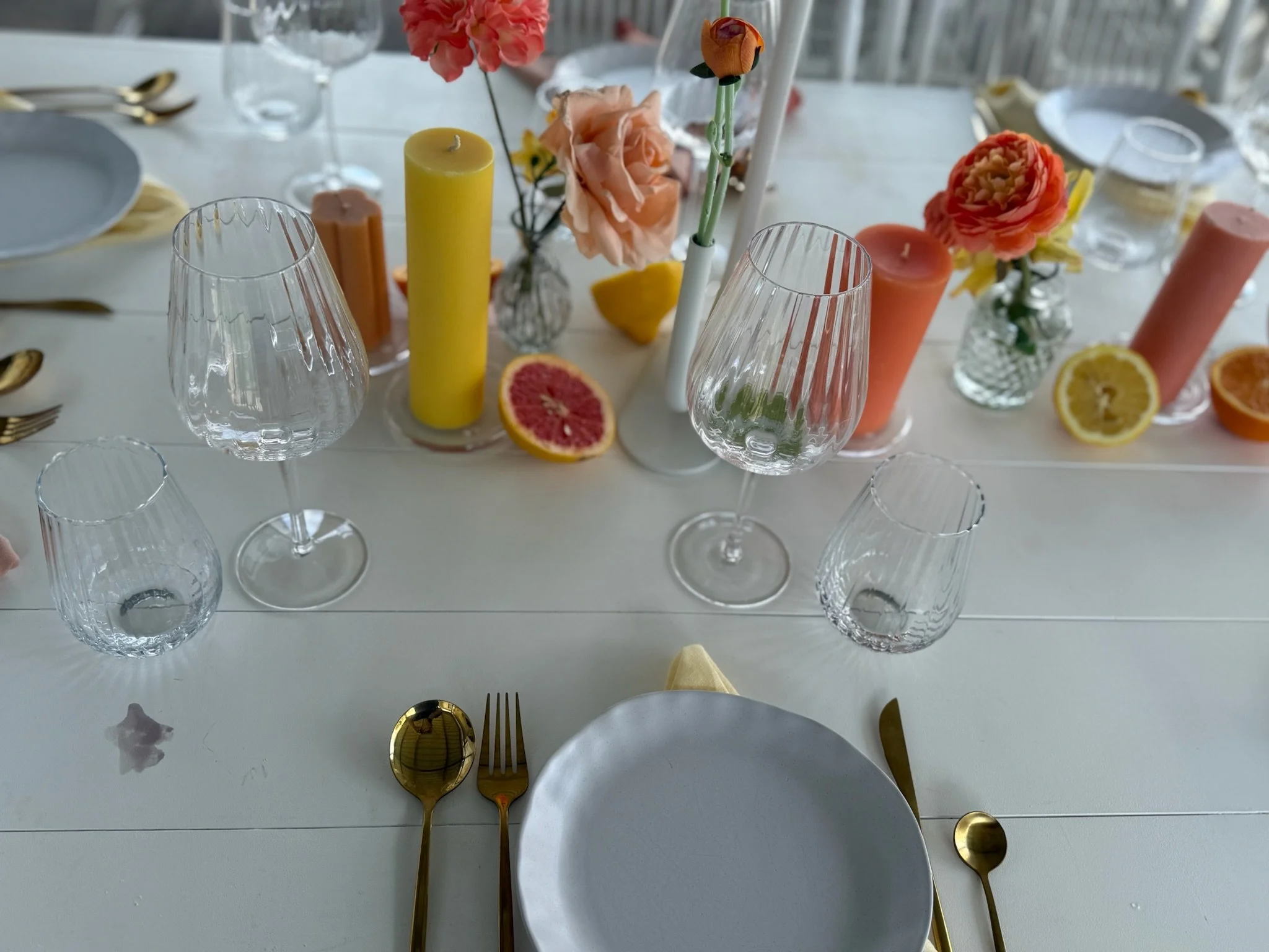 Overhead shot of a decorated table with glassware, gold utensils, white plates, and a centerpiece of pink, orange, and yellow flowers with candles, lemon, and grapefruit slices.