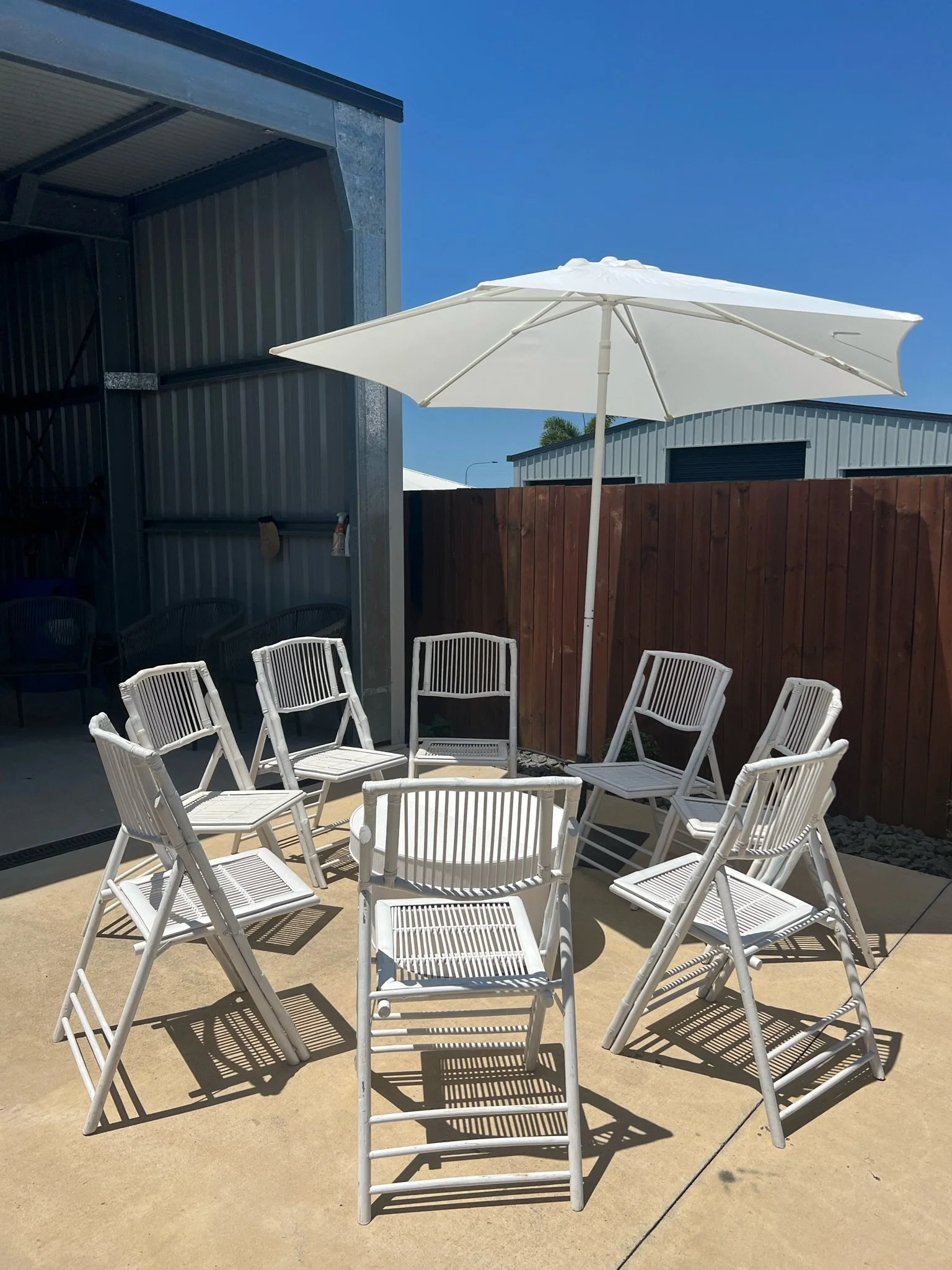 Stacked white outdoor chairs arranged in a circle under a large white patio umbrella on a concrete patio.