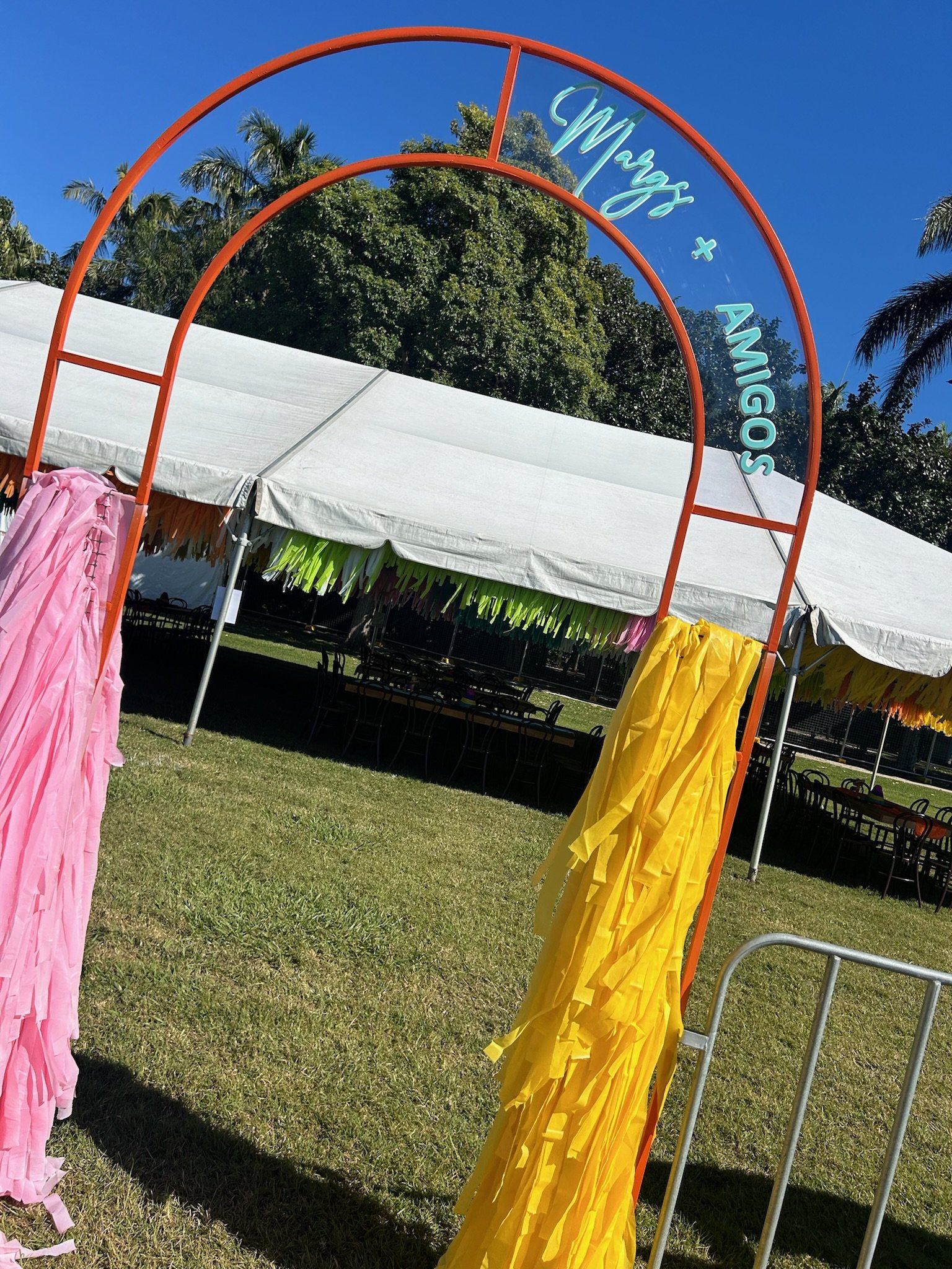 Decorative entrance arch with the words 'Wozz + Amigos' in blue neon sign, pink and yellow tissue paper decorations hanging on either side, outdoor event setup under a white tent with colorful banners, green grass and trees under a clear blue sky.