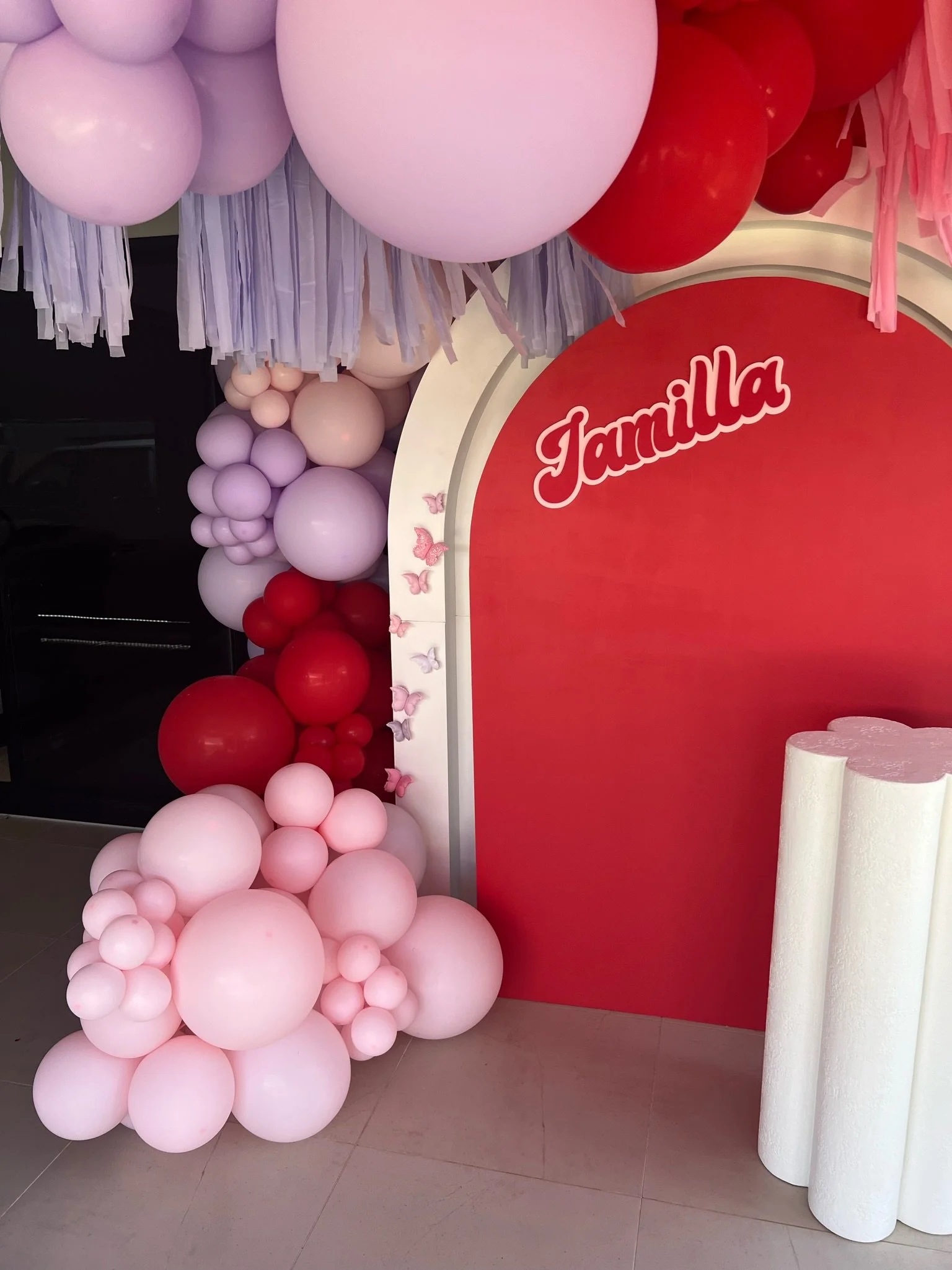 Decorative balloon display with pink, red, and purple balloons, a pink backdrop with the word 'Jamilya' in white script, and white cylindrical foam rolls.