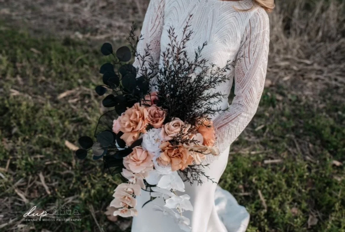 A woman in a long white lace dress holding a bouquet of peach, white, and dark green flowers and foliage outdoors.