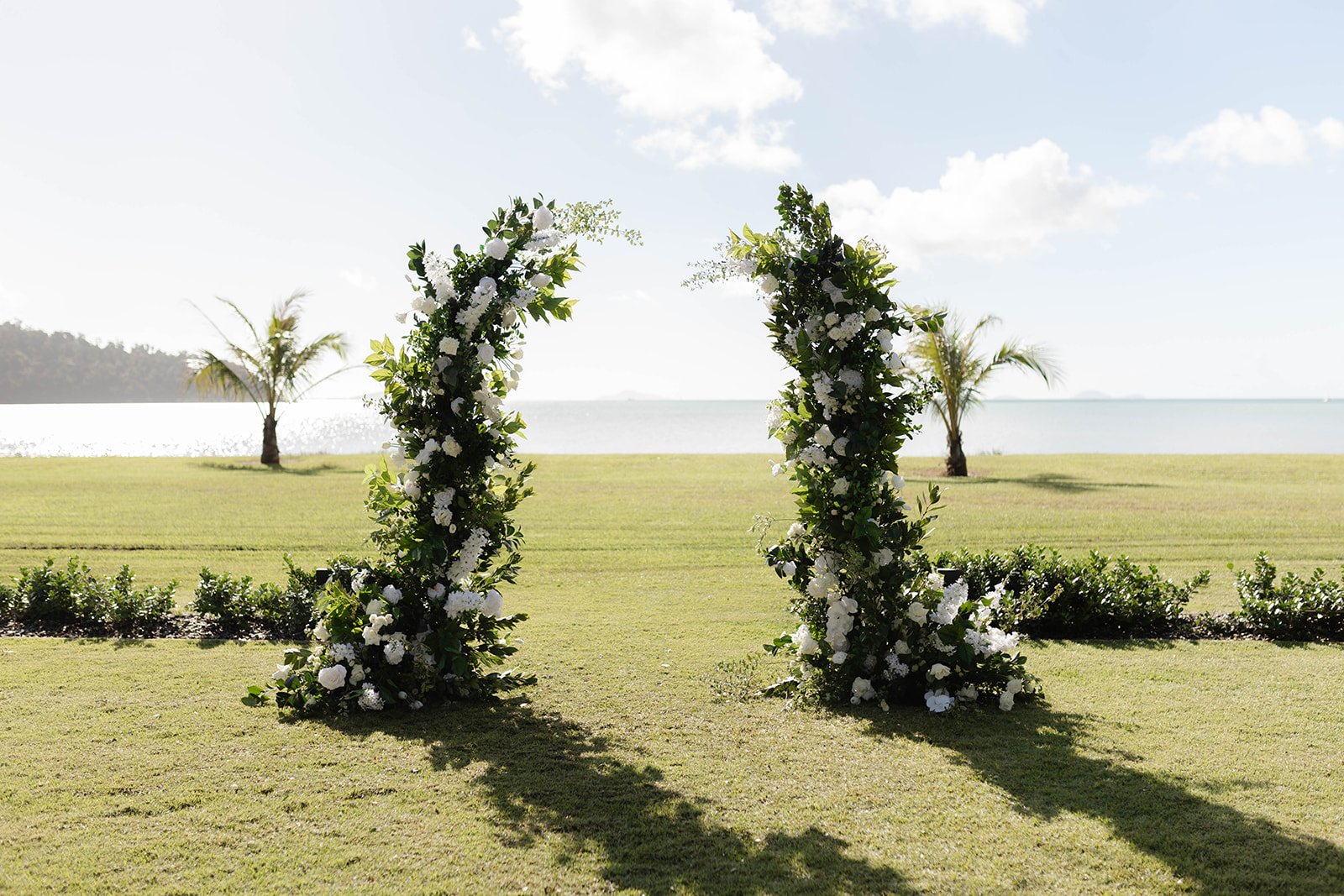 A floral arch decorated with white flowers and greenery on a grassy outdoor wedding venue near the ocean with palm trees in the background.