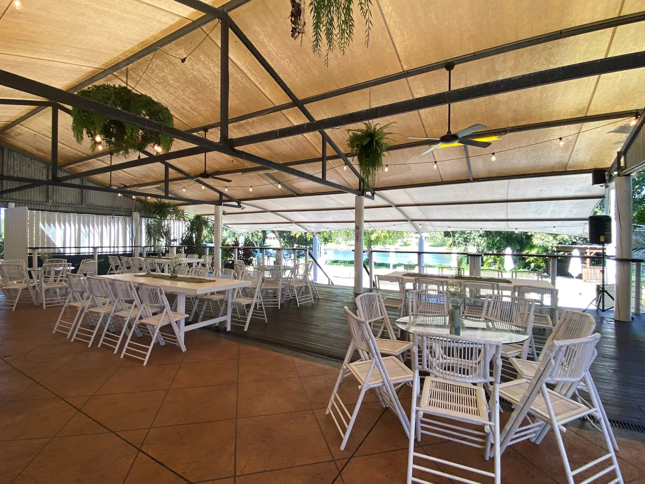 An empty outdoor dining area with white tables and chairs, decorated with potted plants, under a large beige tent with string lights, ceiling fans, and a view of trees and water outside.