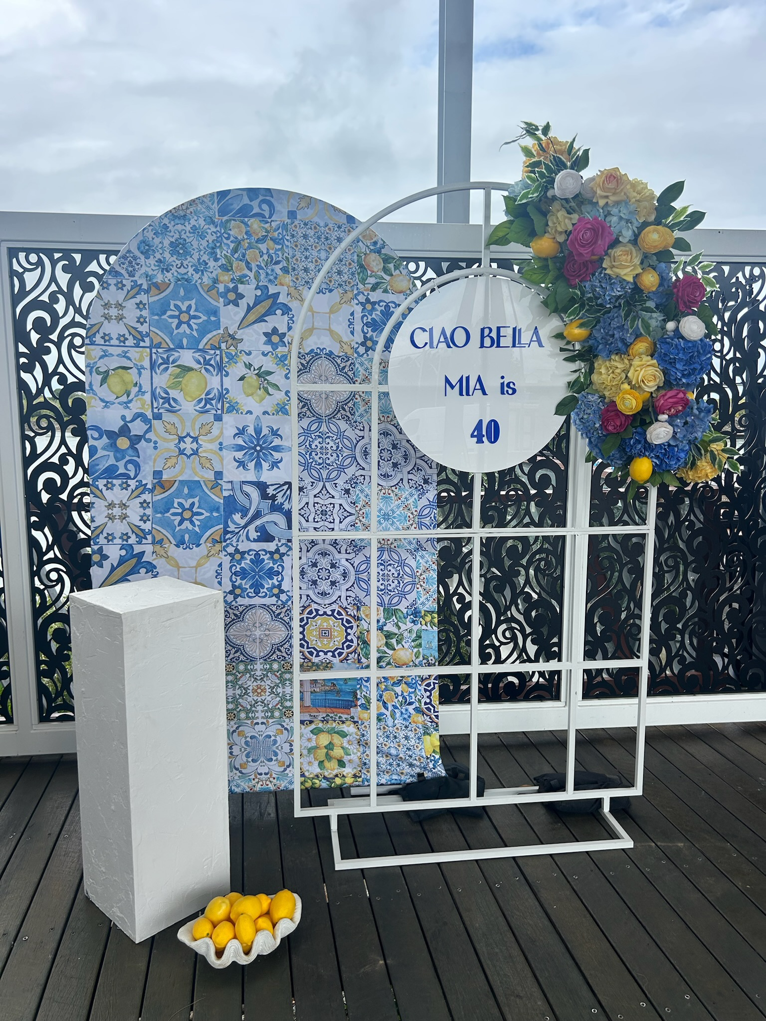 Decorative display with a white circular sign that reads 'CIAO BELLA MIA is 40' hung on a white metal grid frame. The background features blue and white patterned tile panels and a black decorative screen, with a floral arrangement of colorful flower