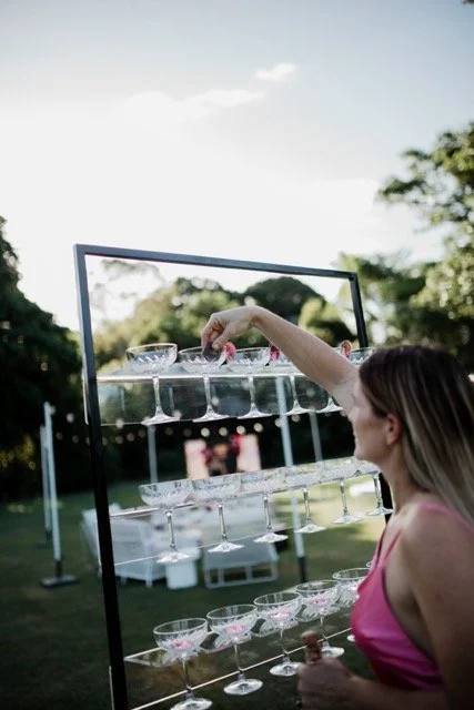 A woman selecting glasses from a large outdoor wine or cocktail glass display set up in a park or garden.