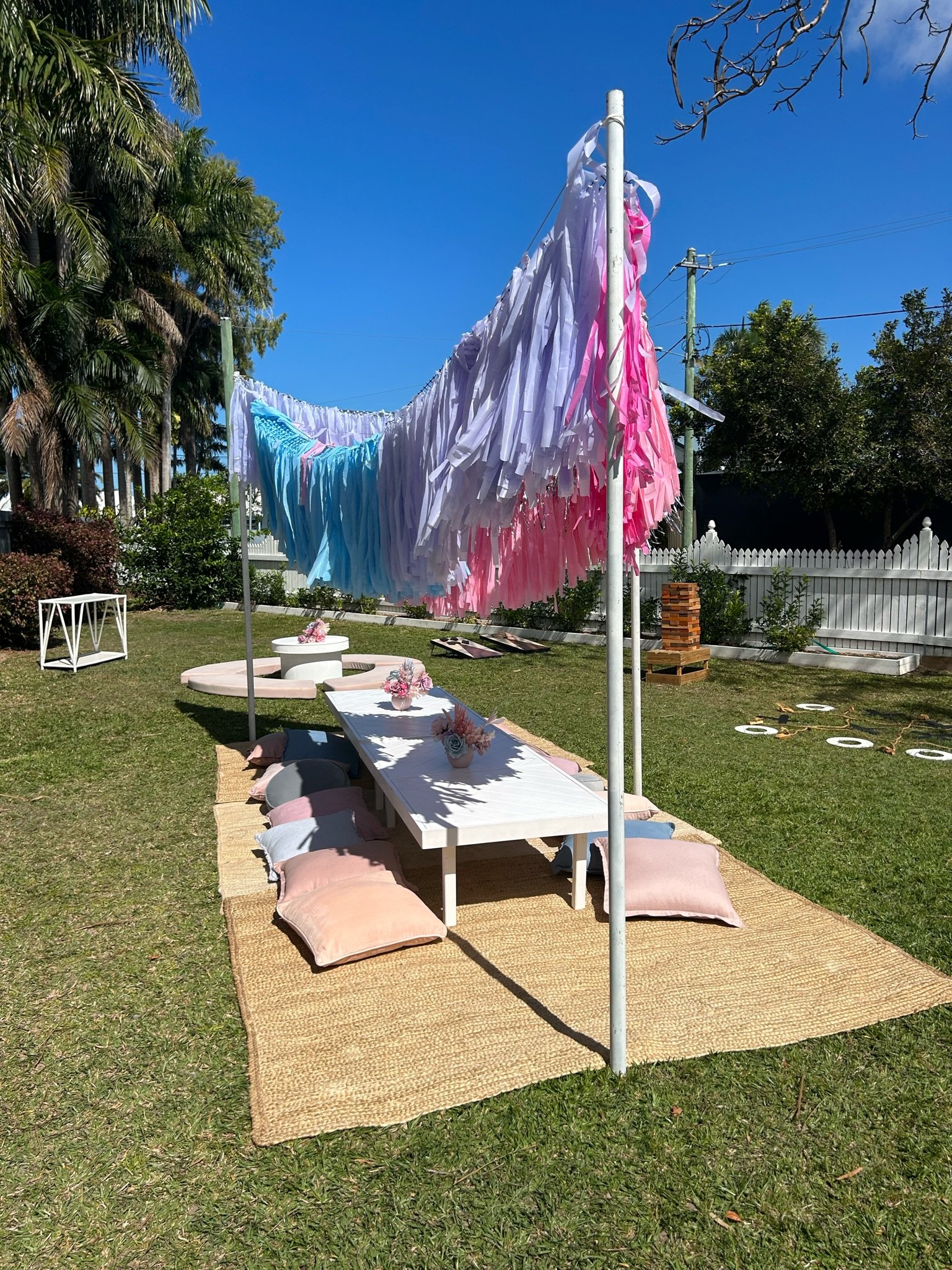 Outdoor setup with a low white table, blush pillows, floral centerpieces, and a colorful tissue paper garland in pink, purple, and blue hanging overhead on a grassy lawn with a white picket fence, trees, and clear blue sky in the background.