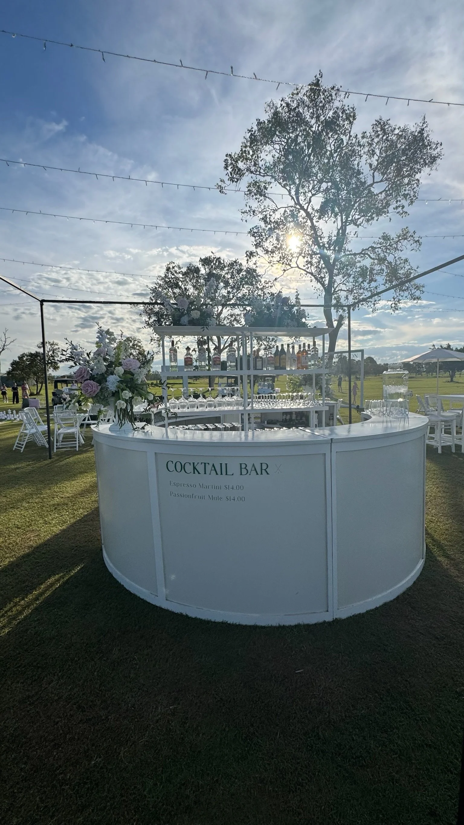White cocktail bar at an outdoor event, decorated with flowers, with a list of drinks and prices. Background shows trees, string lights, and a partly cloudy sky.