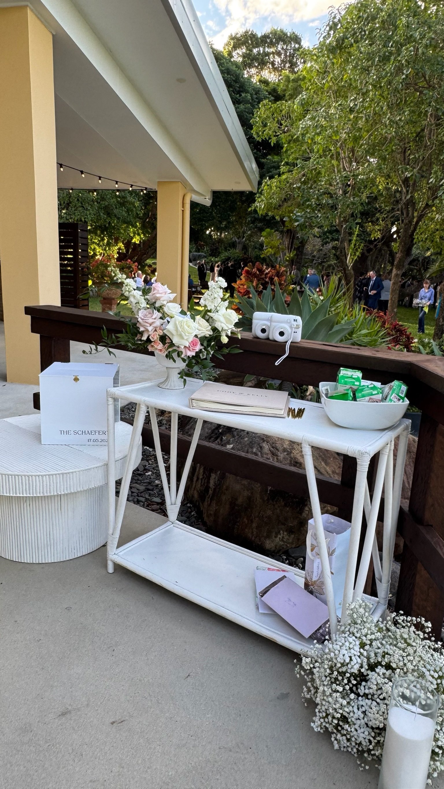 Wedding gift table with flowers, documents, and wrapped gifts outdoors, with guests socializing in the background.