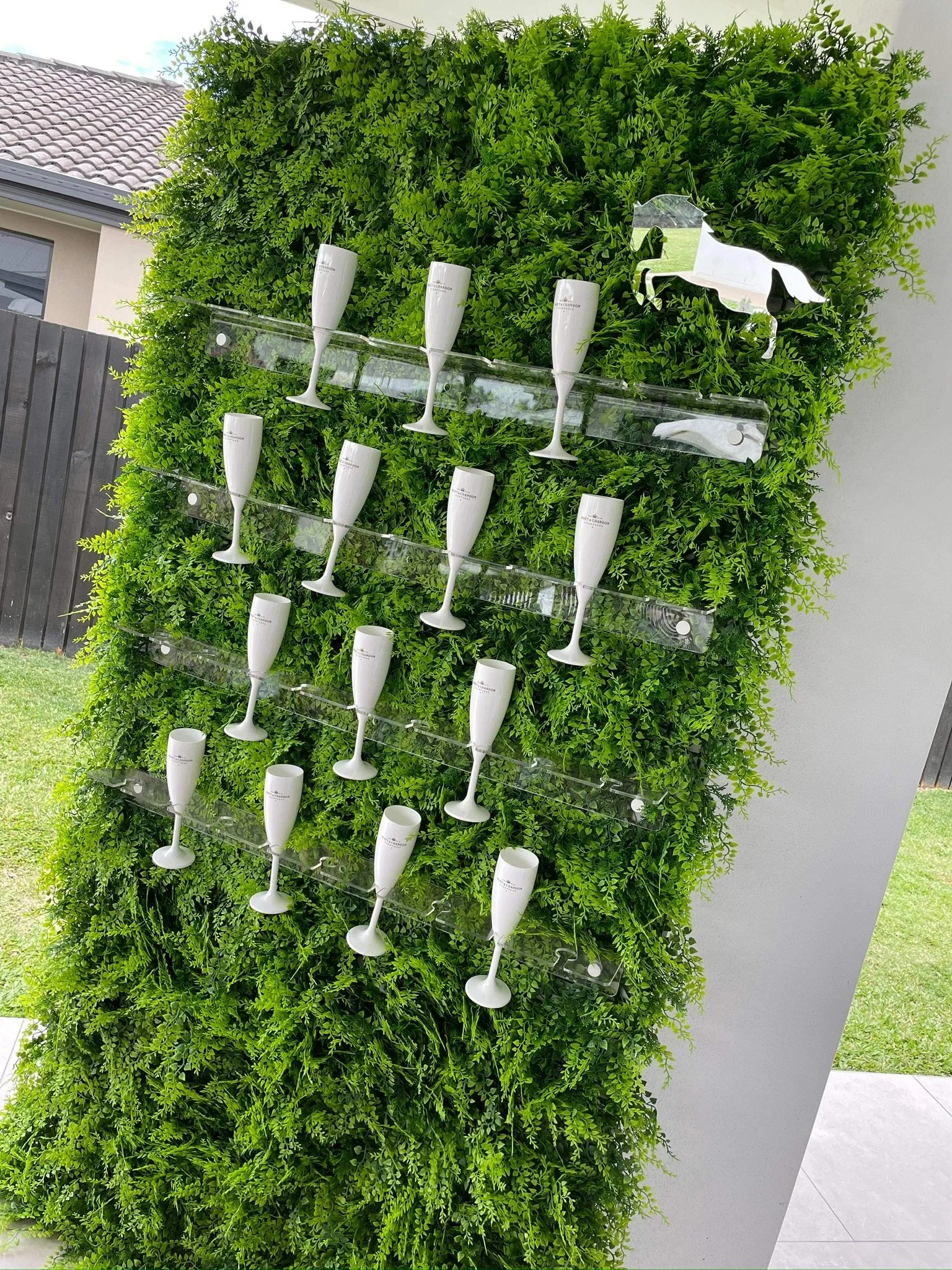 A green vertical garden with 12 champagne flute-shaped glasses mounted on clear acrylic shelves, and a silver horse sculpture on top.