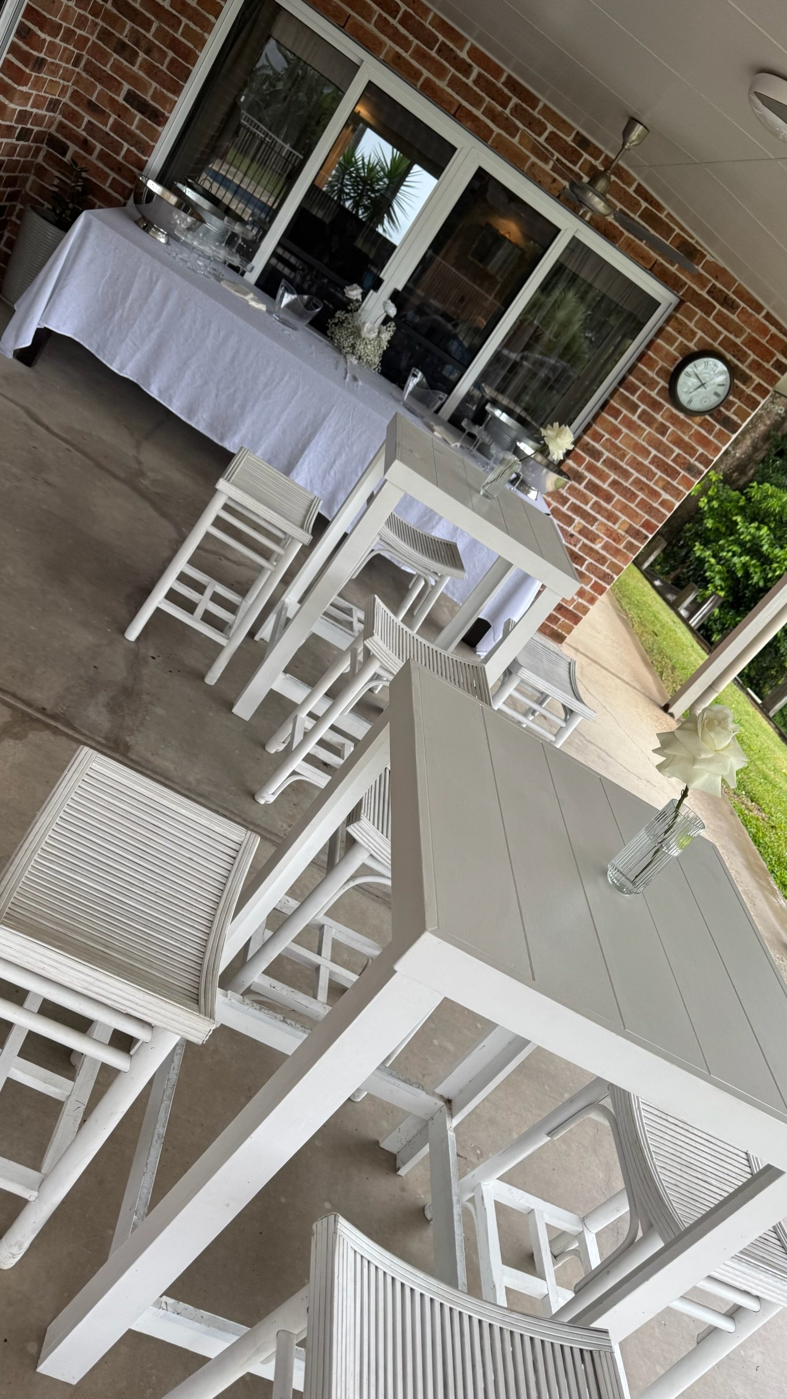 Outdoor patio setup with white tables, chairs, and floral centerpieces, brick wall and sliding glass door in background.