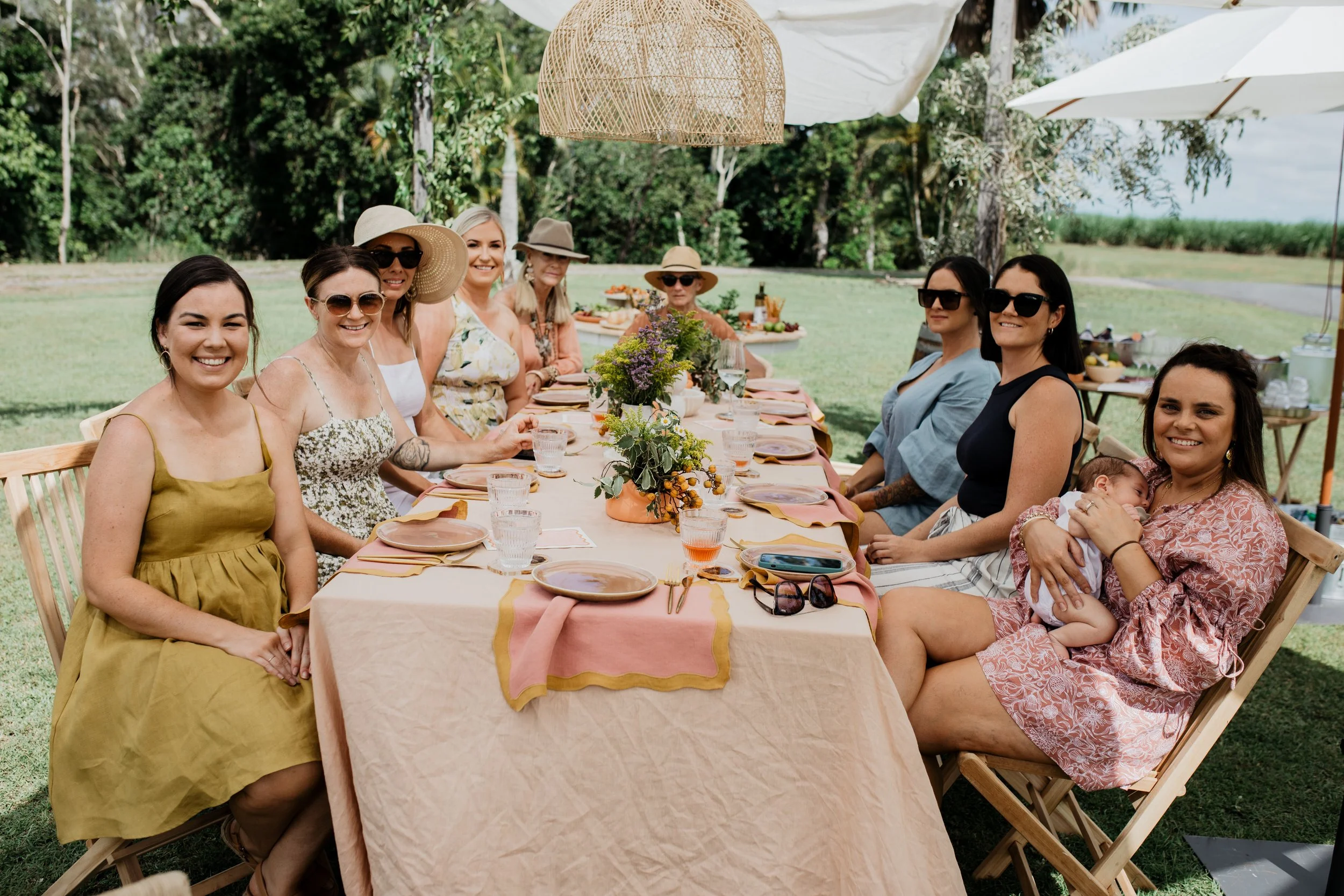 Group of women sitting at a long outdoor table during daytime, enjoying a gathering with greenery in the background.