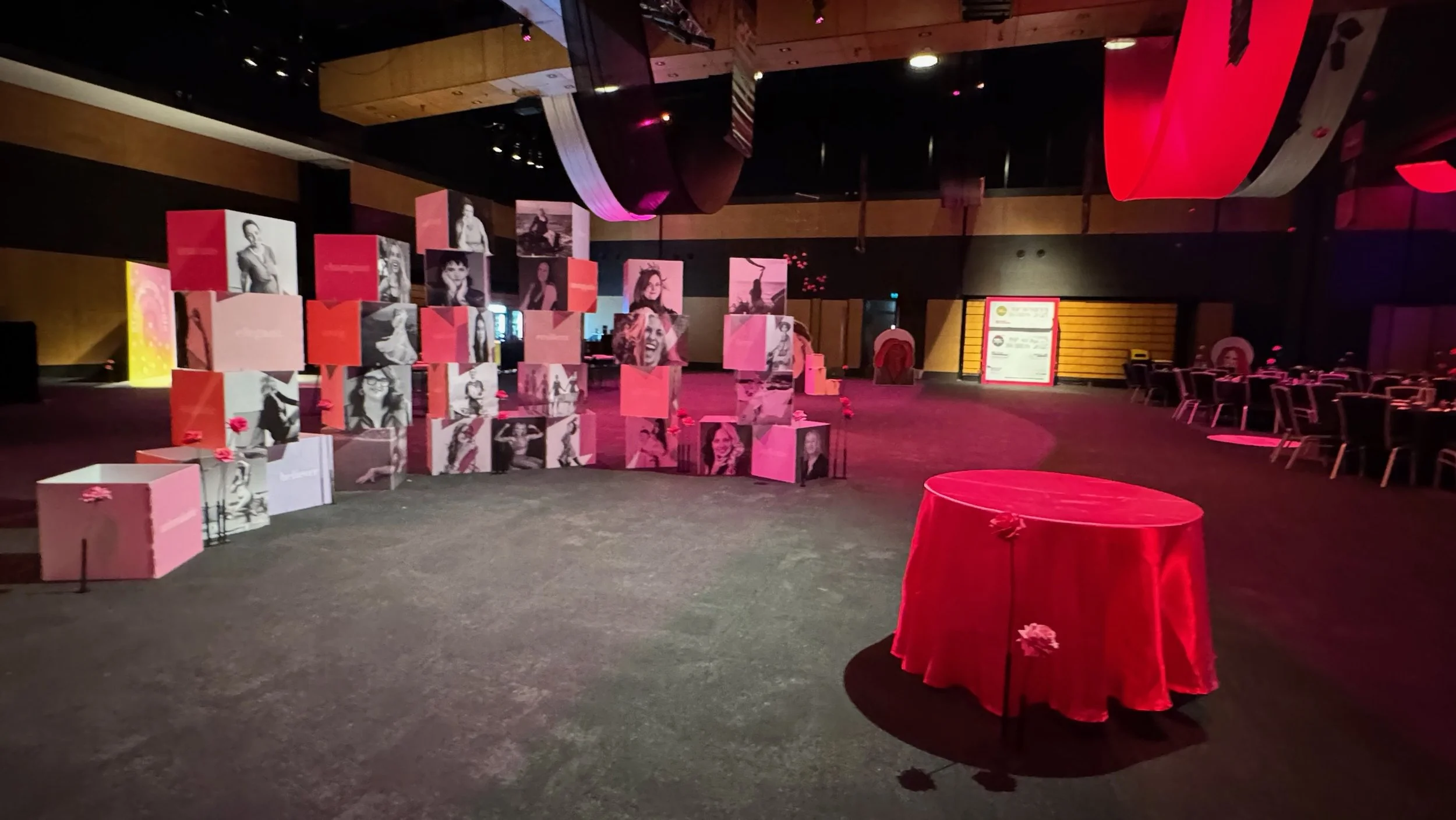Indoor event space decorated with a display of cubes featuring black and white photos of women, pink flowers, and red fabric-covered tables. There are chairs arranged along the sides, and a large framed sign is visible in the background.