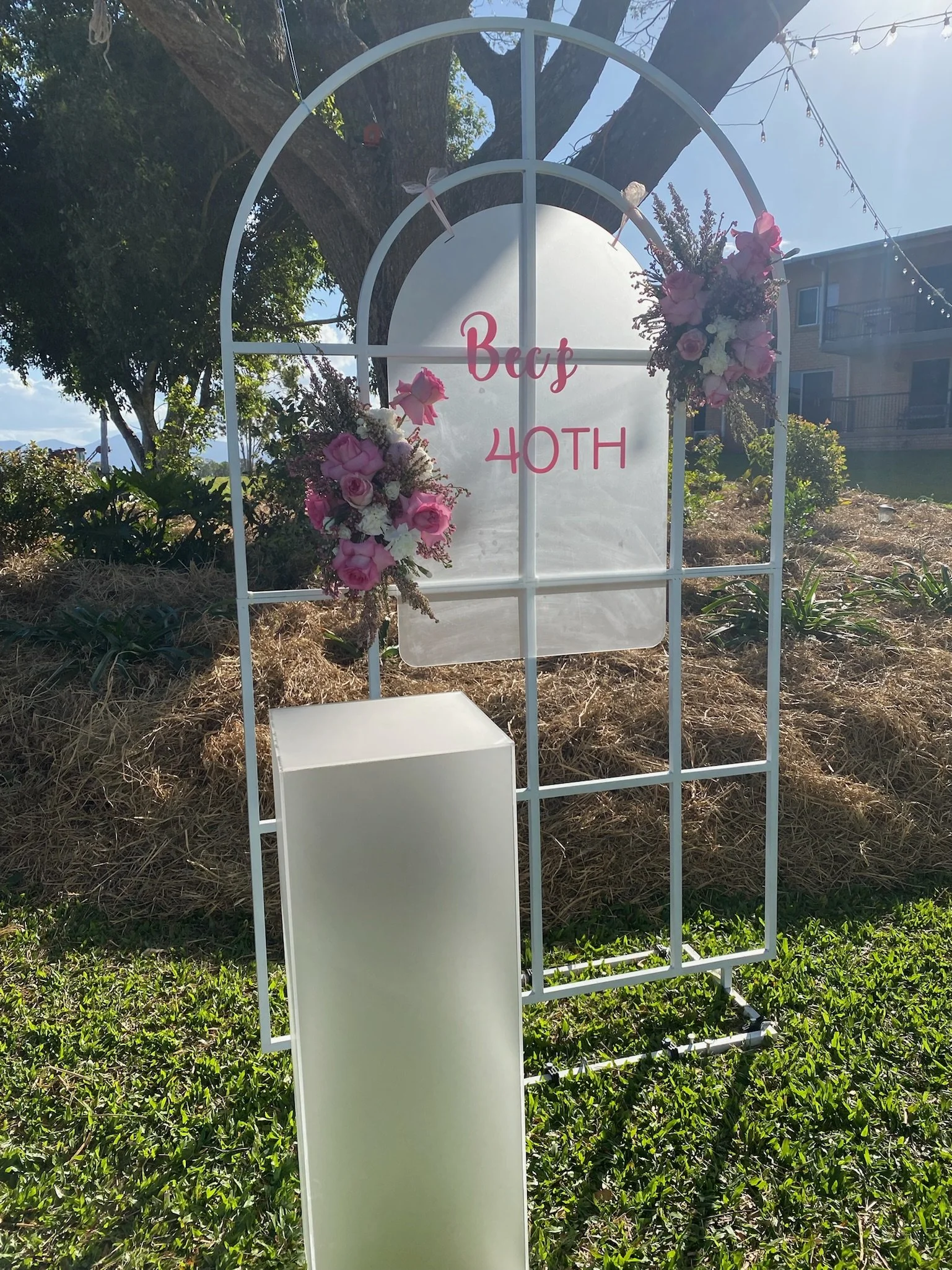 White metal arch decorated with pink and white flowers, with a sign that reads "Becs 40TH" in pink letters, set outdoors with grass and trees.
