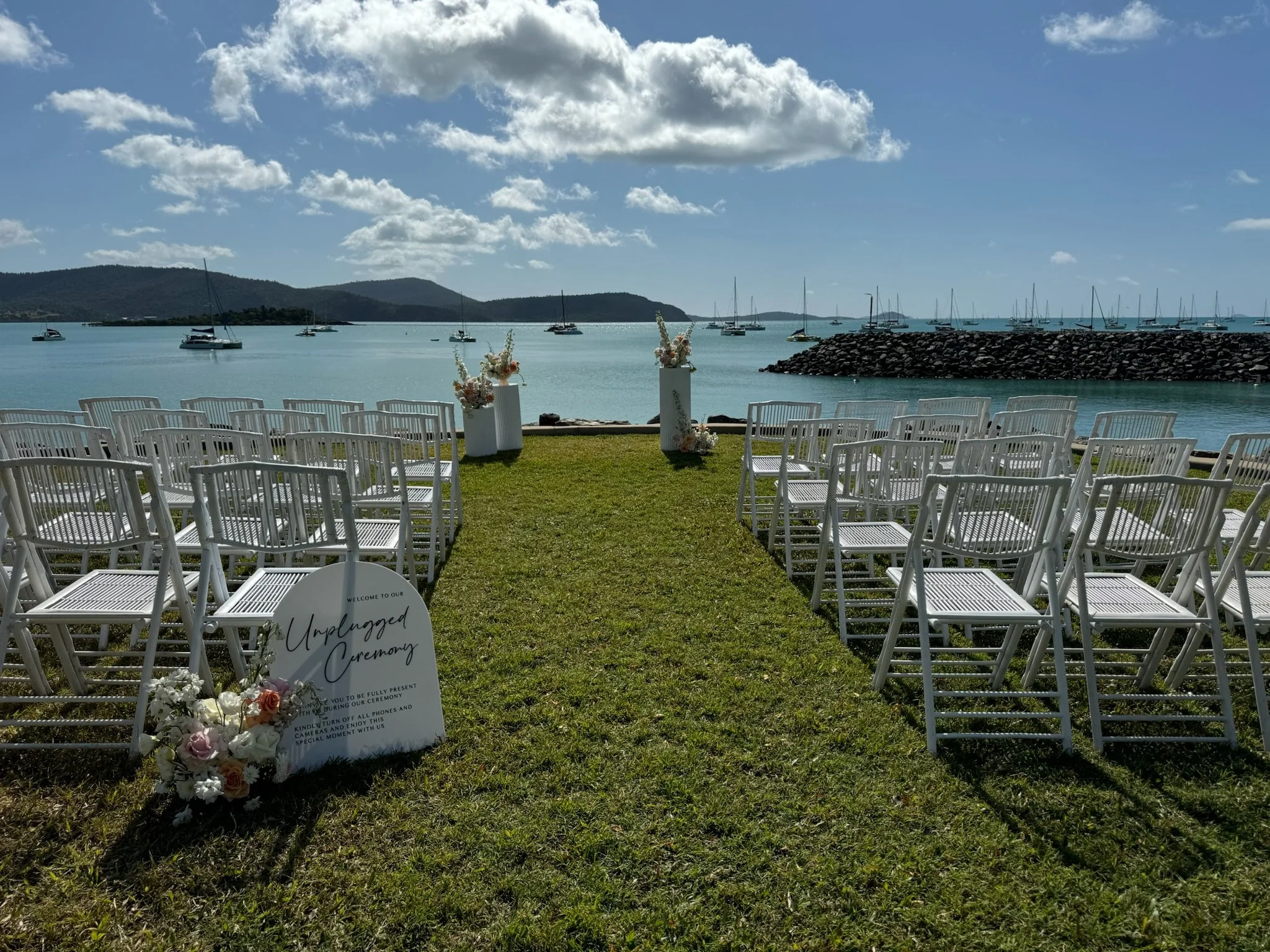 Outdoor wedding ceremony setup by the water with white chairs arranged in rows on grass, floral decorations on white pedestals, and a welcome sign for an unplugged ceremony, with a lake, sailboats, rocky breakwater, and mountains in the background.