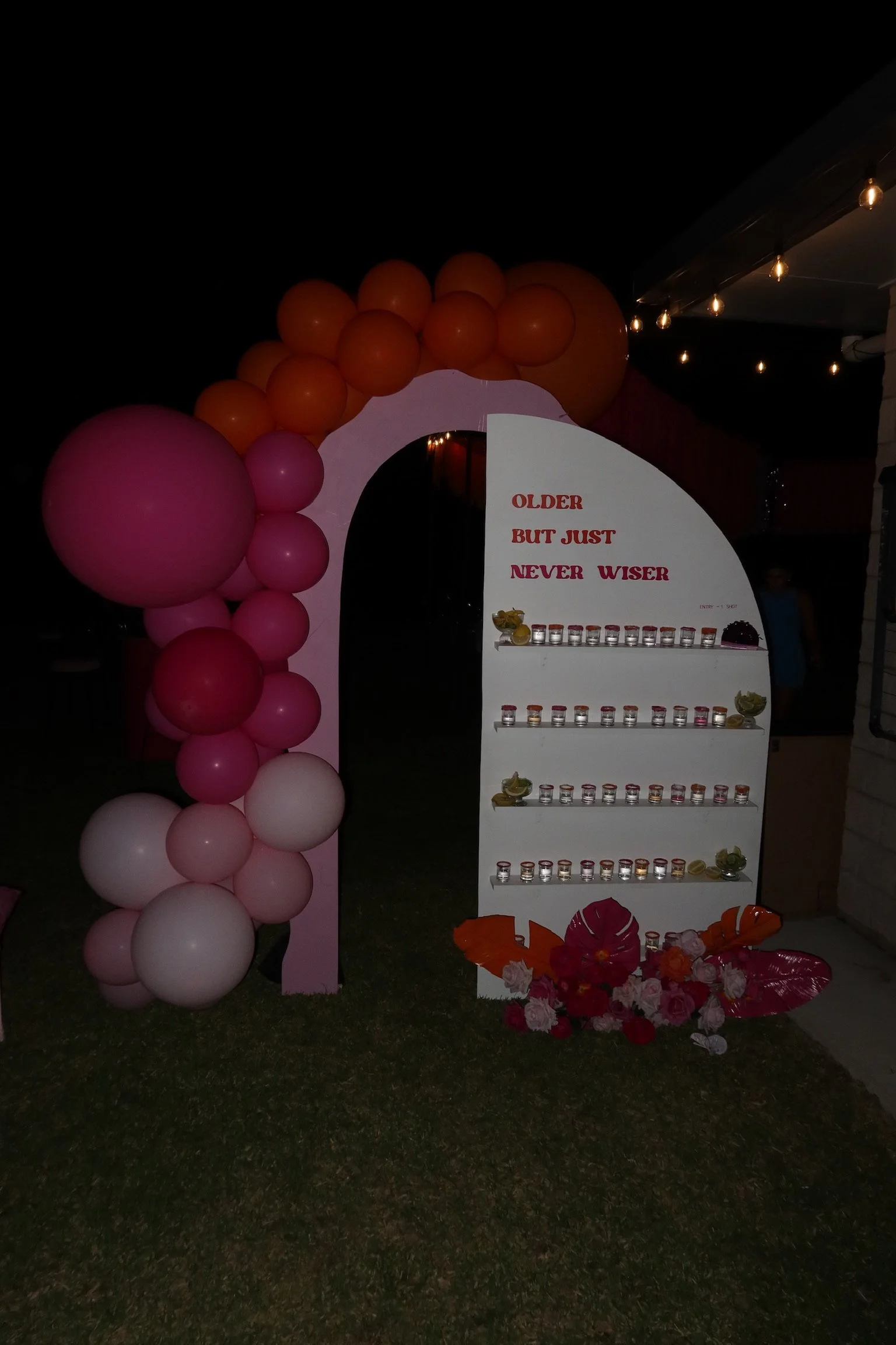 Decorative display with pink and orange balloons, a white stand with shelves holding small containers, pink and red flowers, and a sign stating 'OLDER BUT JUST NEVER WISER' at night.