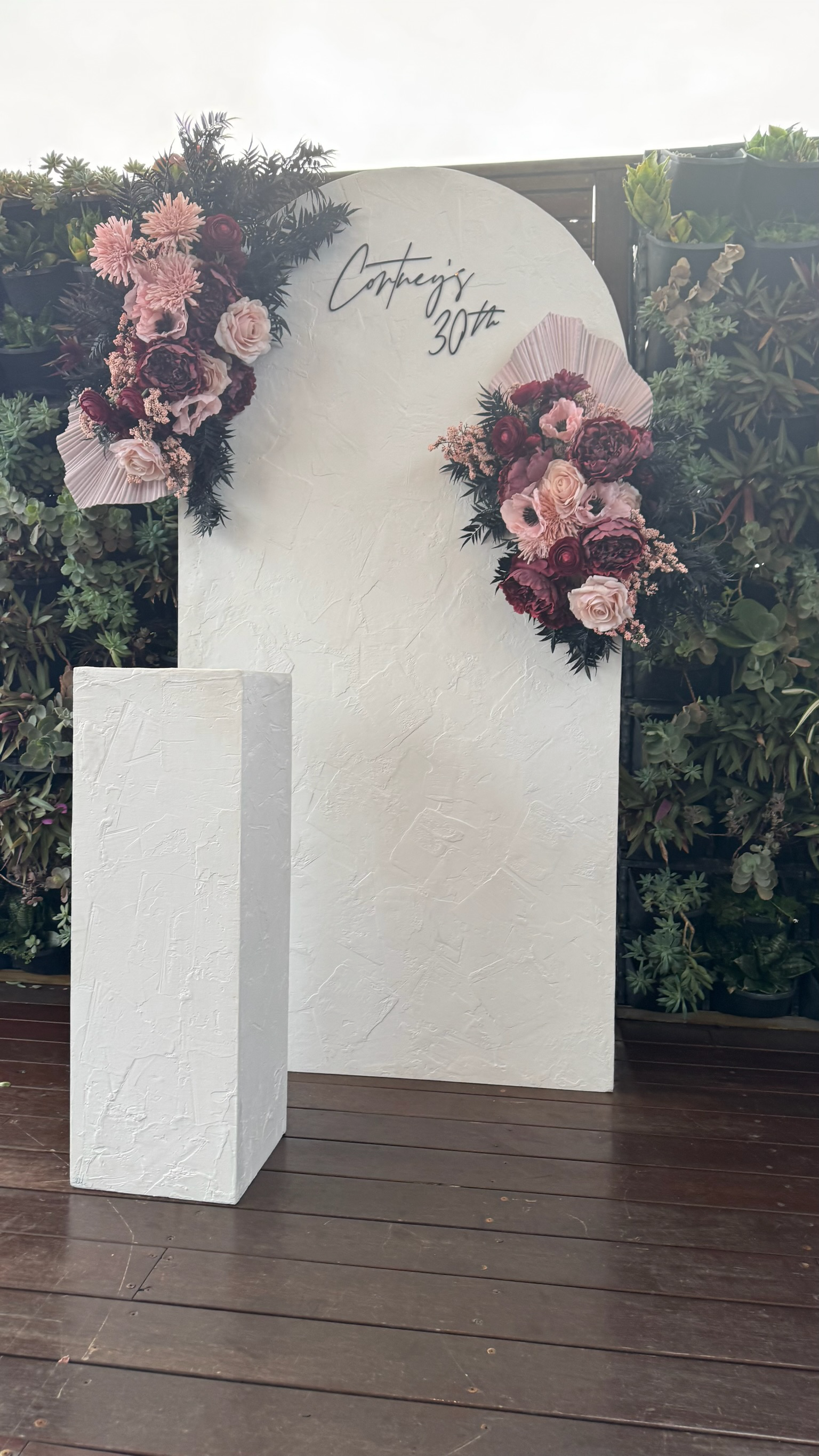 Large white backdrop decorated with pink and red flowers on both sides, with handwritten text reading 'Conting's 30th' at the top. A white pedestal stands in front of the backdrop, all set on a wooden deck with green plants surrounding the setup.