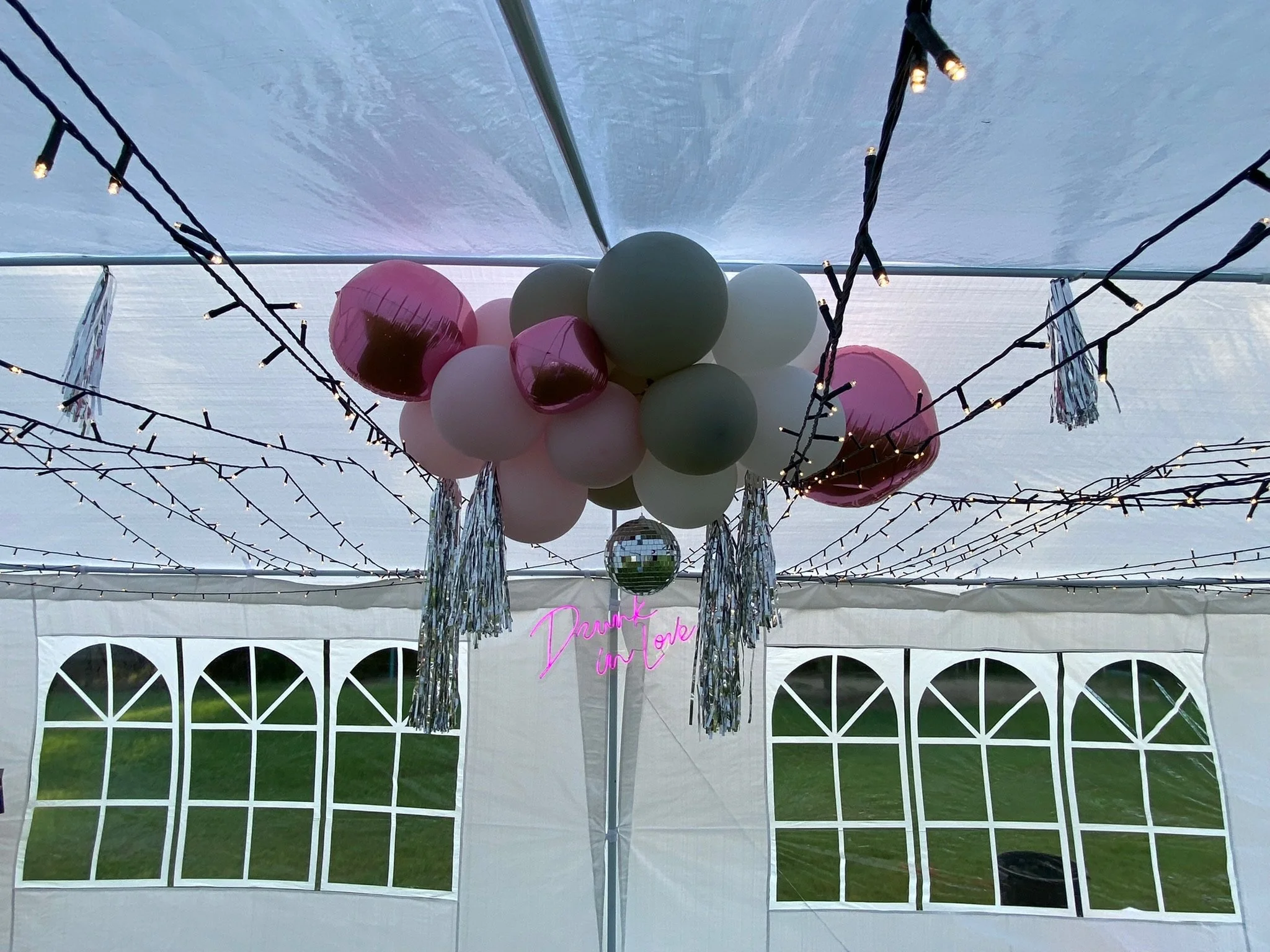 Decorations hanging inside a white tent, including pink, white, and green balloons, metallic fringe tassels, string lights, a mini disco ball, and a pink neon sign that says 'Drink Up'.