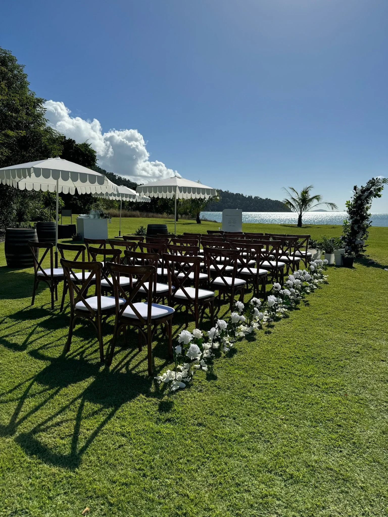 Outdoor wedding ceremony setup with rows of chairs, white flowers lining the aisle, umbrellas, and a view of the water and palm trees in the background on a sunny day.