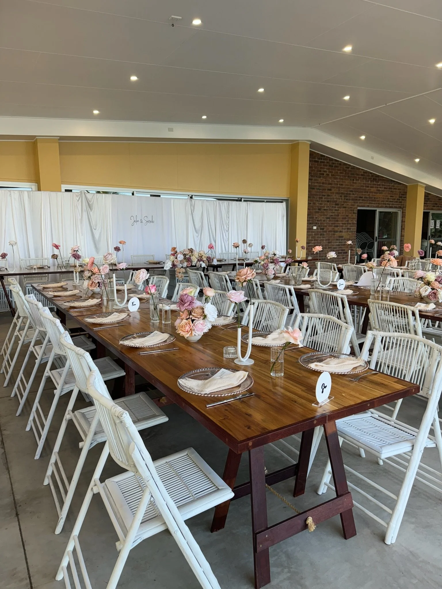 Wedding reception setup with a long wooden table decorated with pink and white flowers, candles, and tableware in a bright indoor space with a white backdrop and brick wall.
