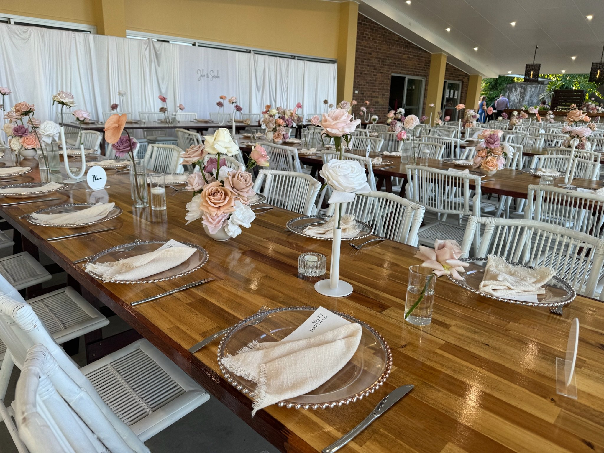 Wedding reception table decorated with pink and white roses in vases, clear glass plates with beige napkins, silverware, clear glass water cups, and a centerpiece with a tall white candle. Additional tables with similar decorations are visible in the